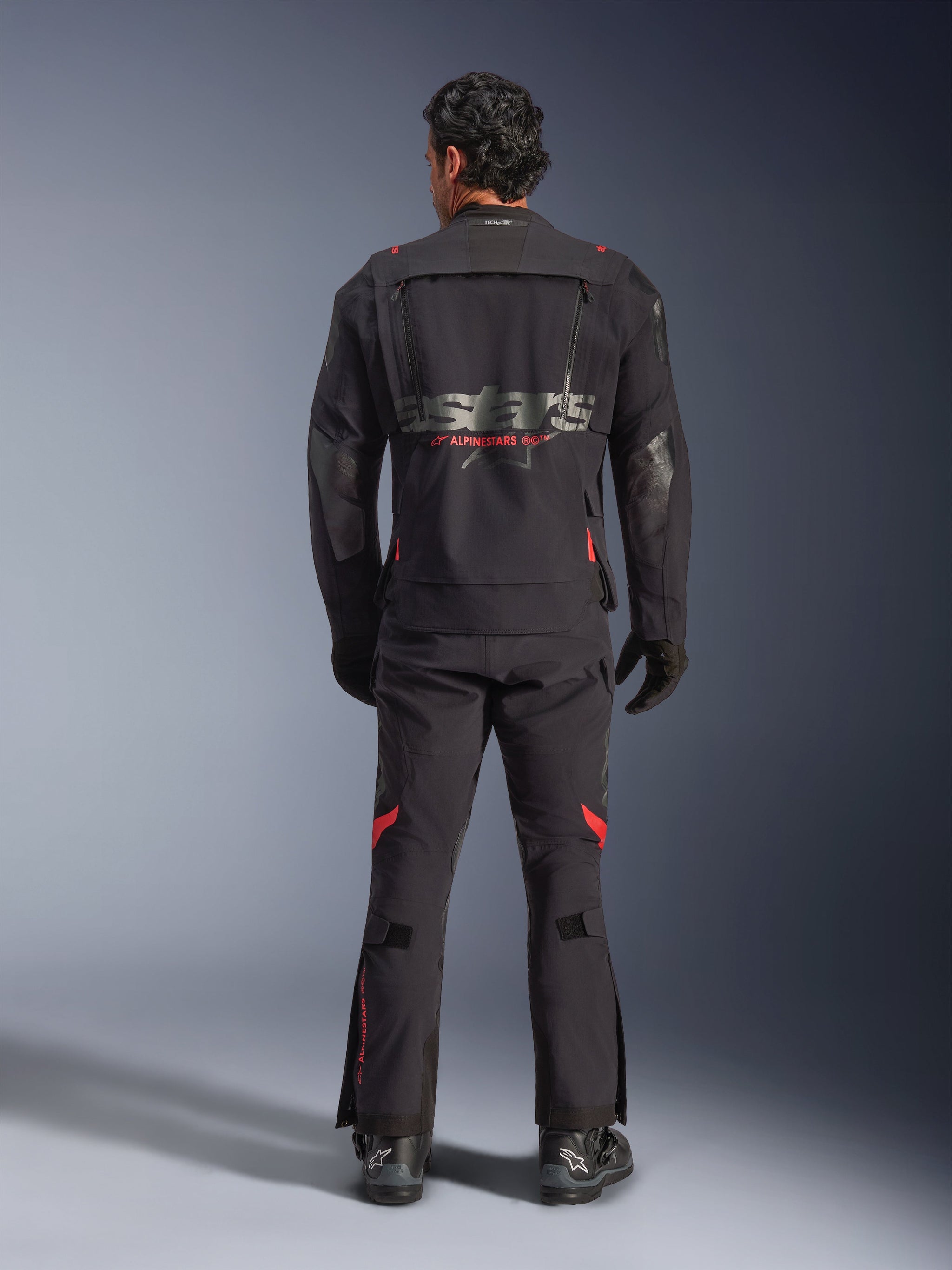 A rider seen from behind wearing the Alpinestars Halo Pro Drystar® XF Laminated Jacket in Black Dark Shadow, featuring a large gray astars logo with red Alpinestars branding, modular ventilation panels on the back, and paired with matching adventure riding pants and black boots.