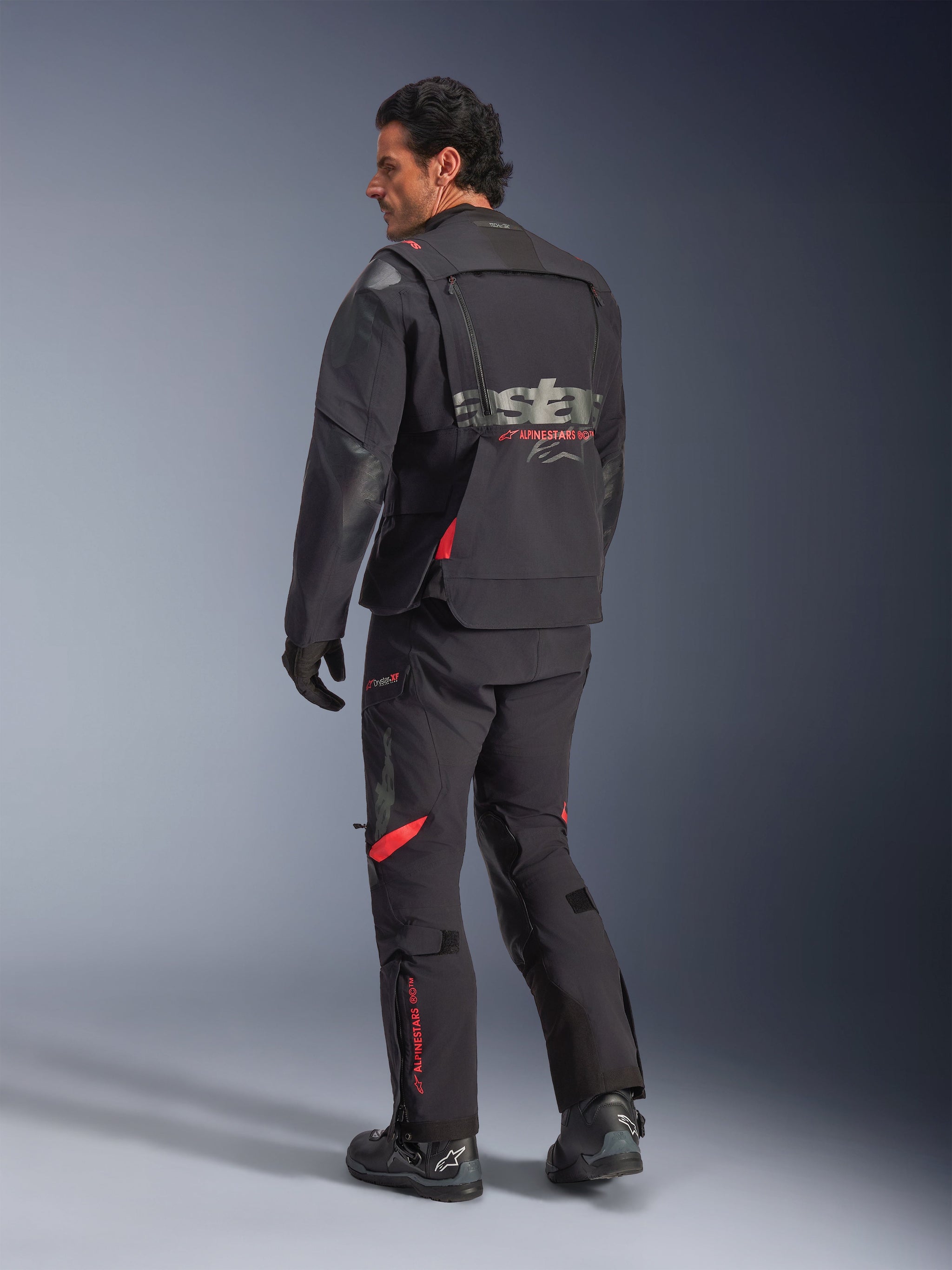 Alpinestars Halo Pro Drystar® XF Laminated Jacket, Adventure Motorcycle Jacket, Black Dark Shadow with red accents, rear view worn with matching pants and boots, featuring grey branding on the back, modular textile design for adventure touring