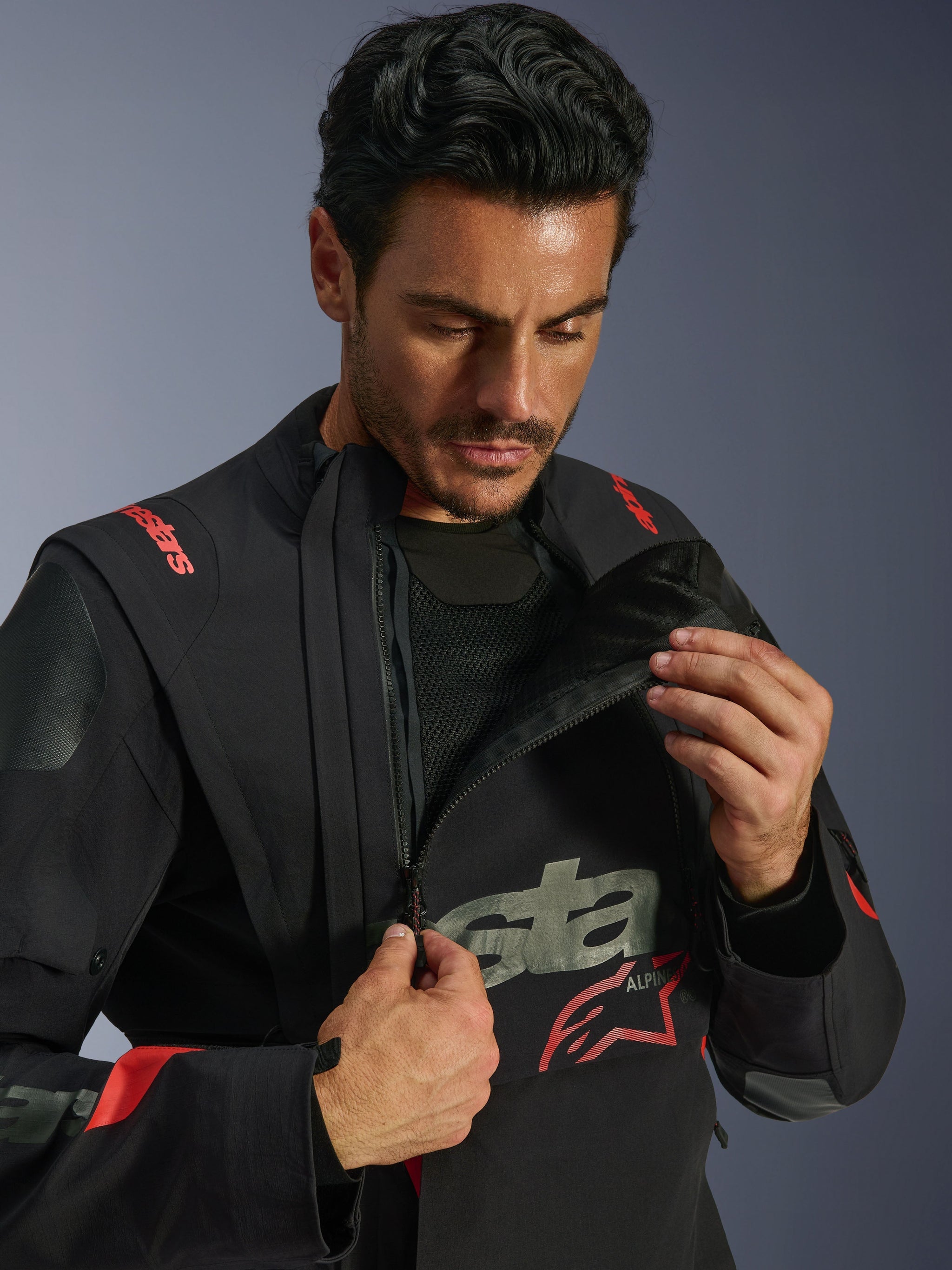 Alpinestars Halo Pro Drystar® XF Laminated Jacket, Black Dark Shadow, model demonstrating the modular ventilation system by unzipping the asymmetrical front chest panel, adventure touring jacket with red Alpinestars branding and technical mesh detailing.