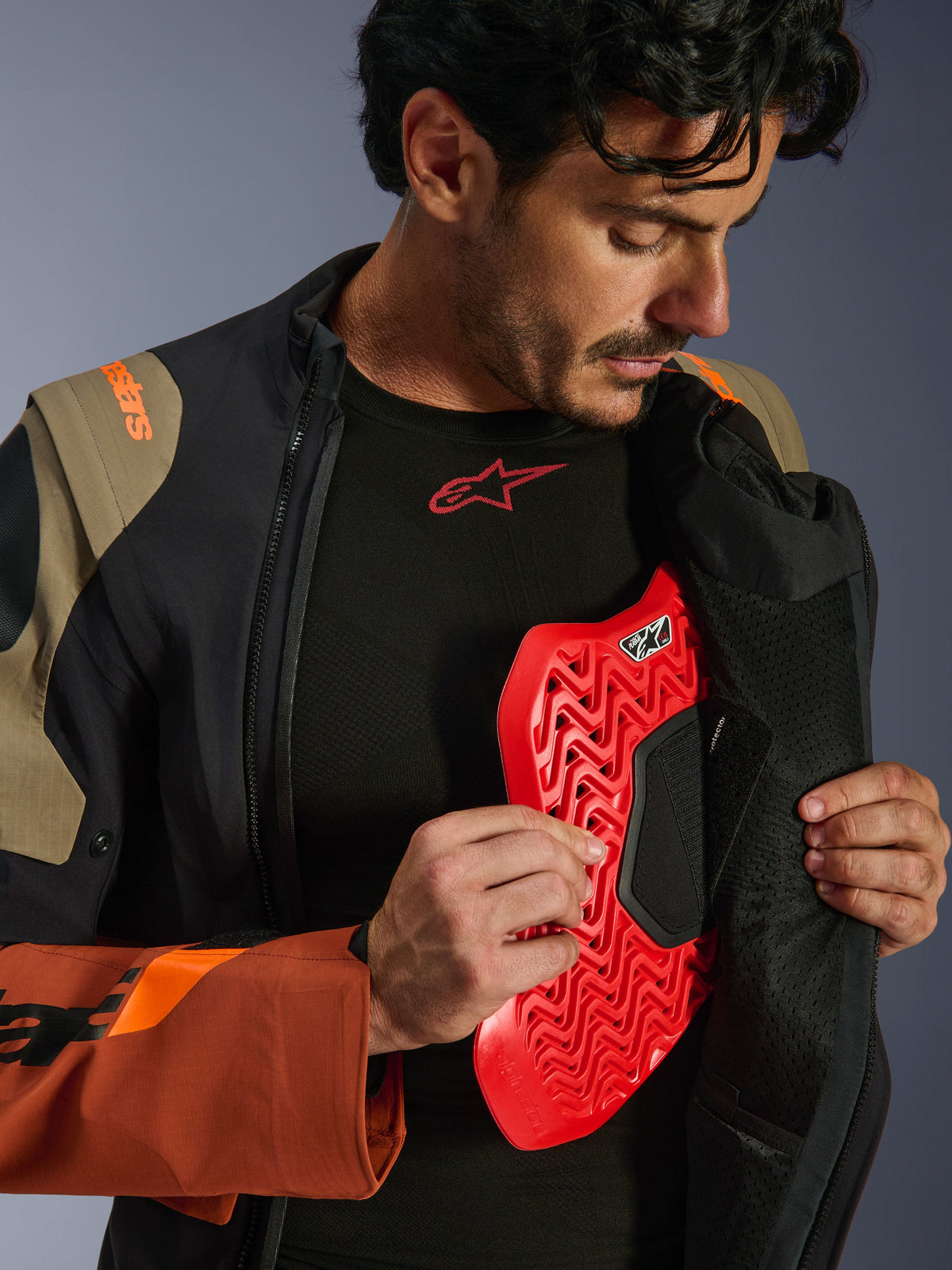Alpinestars Halo Pro Drystar® XF Laminated Jacket in Walnut Rust Black, lifestyle shot of a male model unzipping the adventure jacket to reveal a red chest protector insert, featuring orange branding on the shoulder and a black base layer with a red logo.