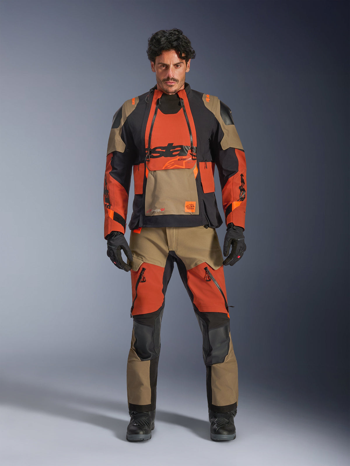 Male model wearing Alpinestars Halo Pro Drystar® XF Laminated Jacket and matching pants in Walnut Rust Black, modular adventure motorcycle gear featuring front ventilation panels and zippers, styled with black motorcycle gloves and boots.