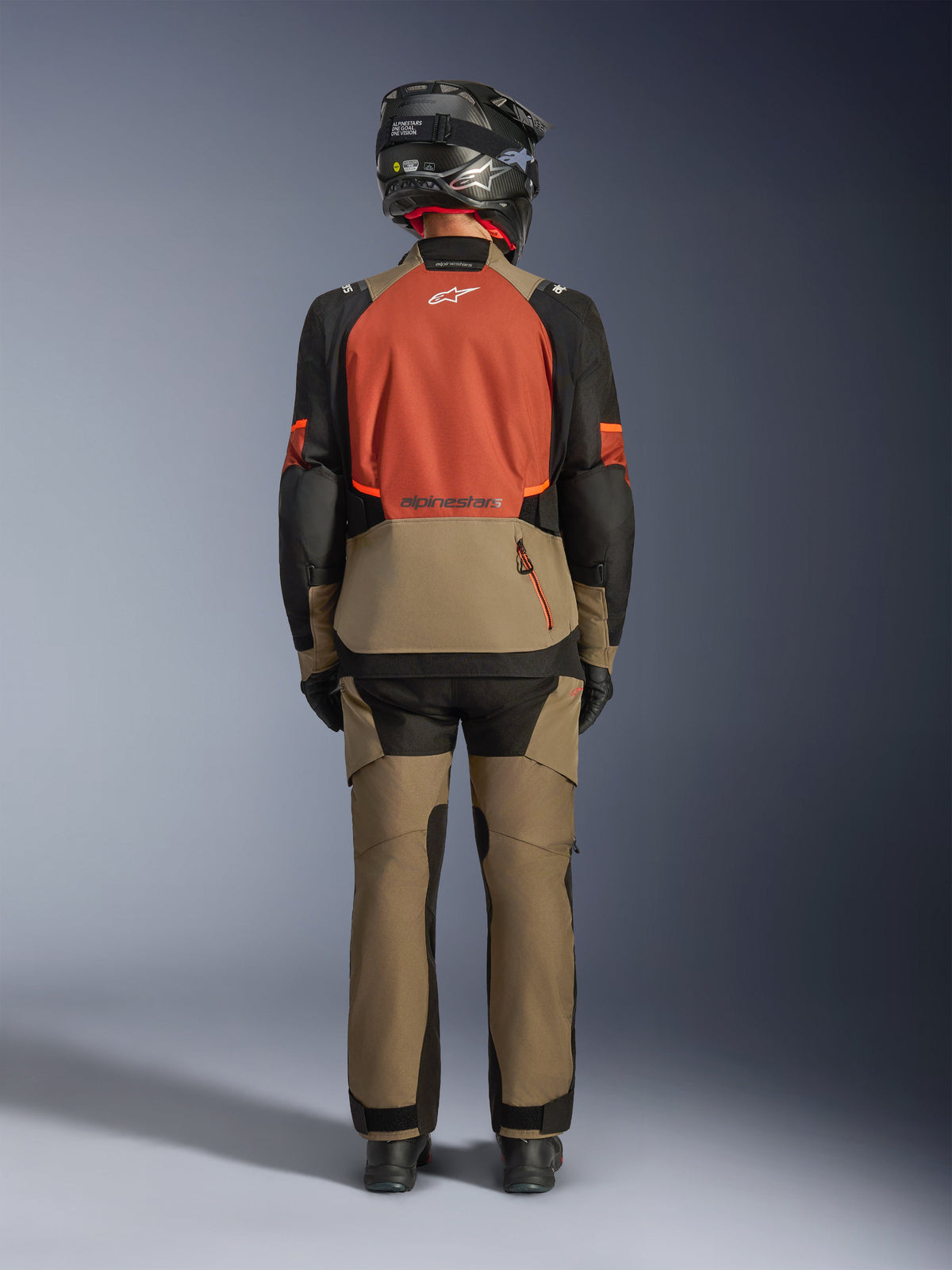 Rear view of a rider wearing the Alpinestars Andes V4 Drystar® Jacket in Walnut Rust Black, paired with matching adventure pants and a black helmet, showcasing the jacket's rust-colored back panel, large rear storage pocket, and reflective logos.