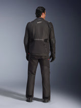 Rear view of a male rider wearing the Alpinestars Andes V4 Drystar® Jacket in Black, paired with matching motorcycle pants and gloves, featuring the Alpinestars logo and wordmark on the back, technical all-weather adventure gear