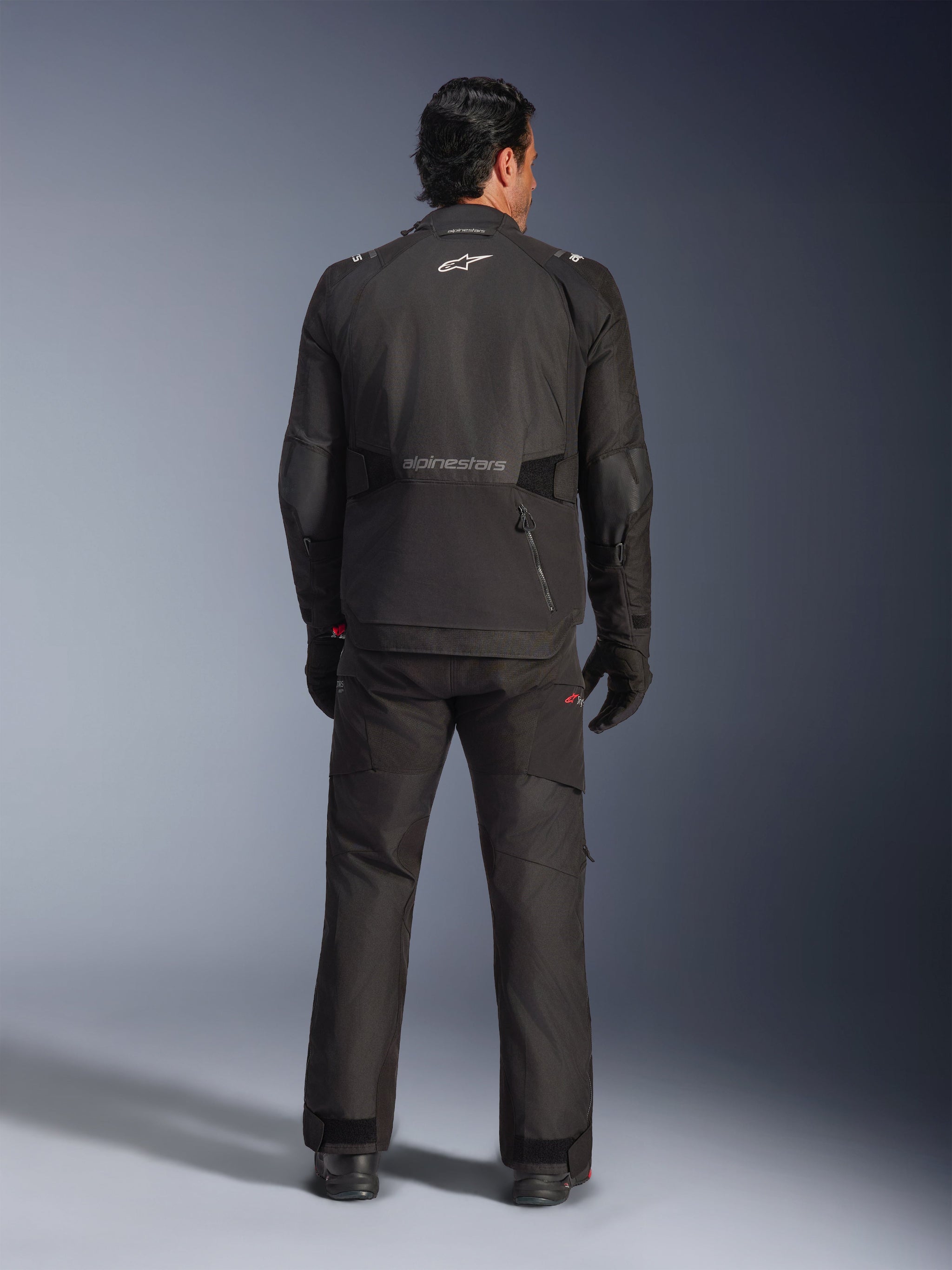 Rear view of a male rider wearing the Alpinestars Andes V4 Drystar® Jacket in Black, paired with matching motorcycle pants and gloves, featuring the Alpinestars logo and wordmark on the back, technical all-weather adventure gear