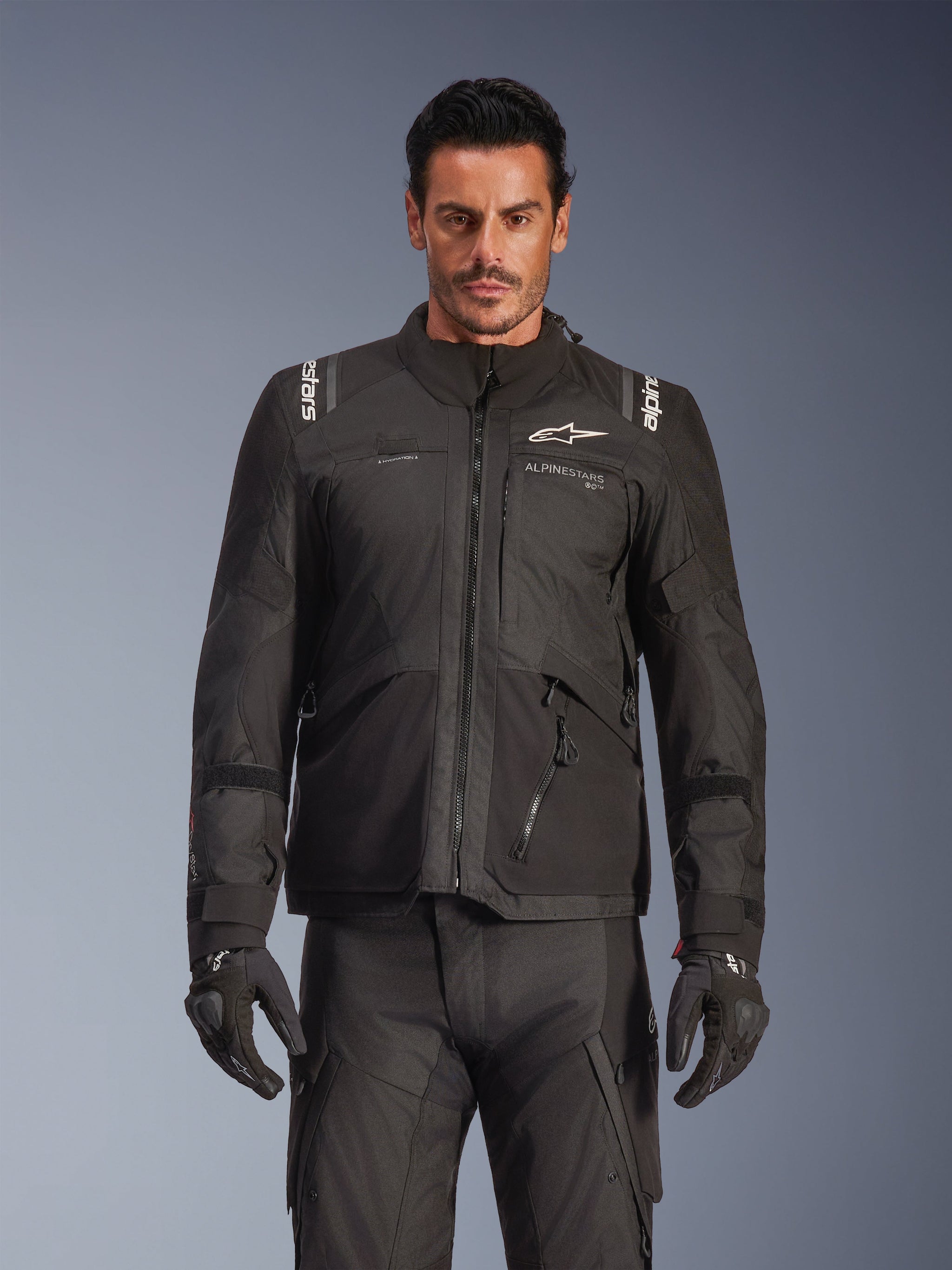 Male model wearing the Alpinestars Andes V4 Drystar® Jacket in Black, adventure touring motorcycle jacket, paired with matching black riding pants and gloves, featuring waterproof zippers and white Alpinestars branding on the chest and shoulders, studio shot for all-weather riding gear.