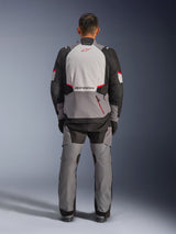 Alpinestars Andes V4 Drystar® Jacket, Adventure Motorcycle Jacket, Dark Gray Ice Gray Black and Bright Red, rear view of a rider wearing matching pants, featuring a large ice gray back panel with red logo, black reinforced shoulder sections, and adjustable waist straps for all-weather touring.