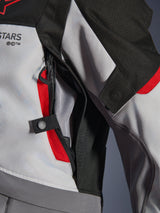 Close-up detail of the Alpinestars Andes V4 Drystar® Jacket in Dark Gray, Ice Gray, and Bright Red, highlighting a zippered chest ventilation port with mesh lining and a snap button adjustment strap.