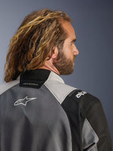 Close-up back profile of a man with long wavy hair wearing the Alpinestars Andes V4 Drystar® Jacket in Dark Gray and Ice Gray, featuring textile panels and a white Alpinestars logo on the back, adventure motorcycle apparel