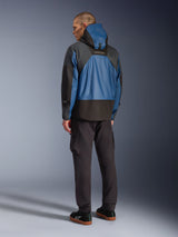 Rear view of a male model wearing the Alpinestars Mospher Waterproof Jacket in Black and Dark Petrol, featuring a hooded design and tactical ripstop construction, paired with black cargo pants and sneakers.