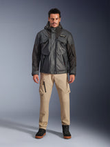 Male model wearing the Alpinestars Mospher Waterproof Jacket in Black, urban contemporary motorcycle gear, tactical utility design with multiple functional pockets and hood, paired with tan cargo pants and black shoes, full-body studio portrait.