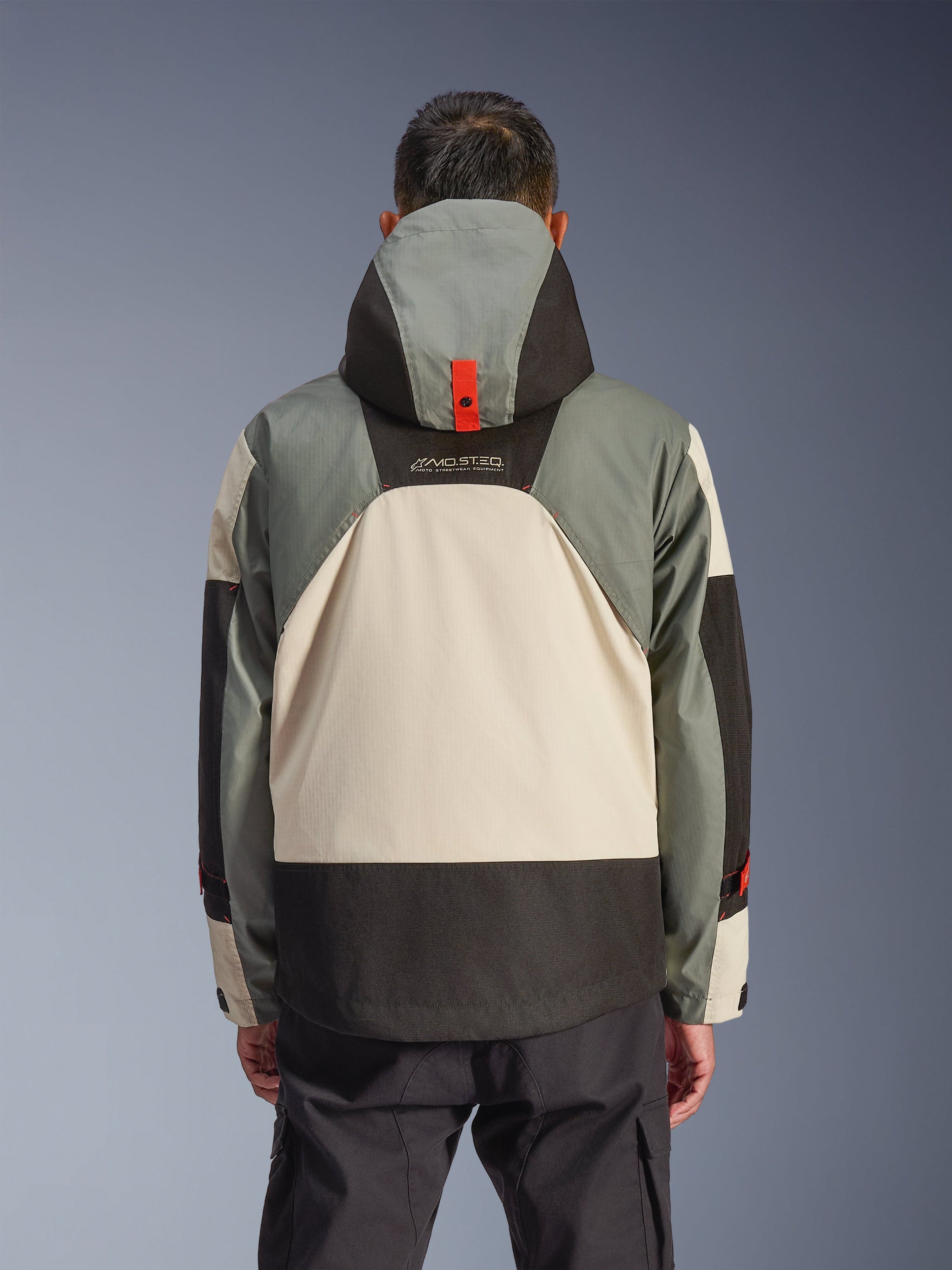 Rear view of the Alpinestars Mospher Waterproof Jacket in Fur Gray Black Ice, featuring a multi-tonal gray, cream, and black panel design, hooded parka silhouette with red snap anchoring detail, 'MO.ST.EQ.' branding, paired with black cargo riding pants