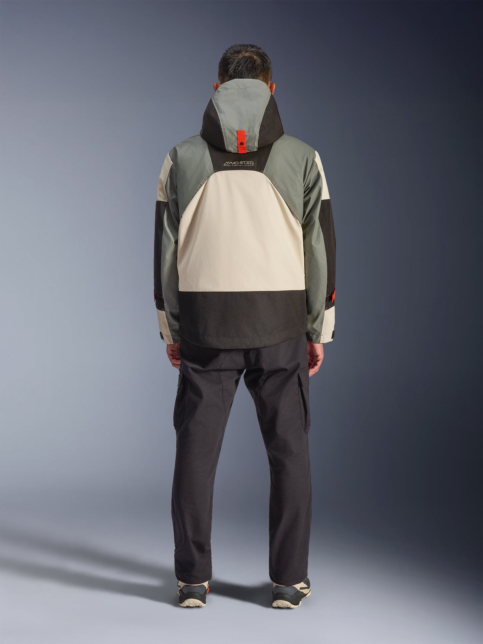 Rear view of a rider wearing the Alpinestars Mospher Waterproof Jacket in Fur Gray Black Ice, tactical-inspired parka with gray, black, and cream color-blocking, features a hood with orange strap detail, paired with black cargo pants.