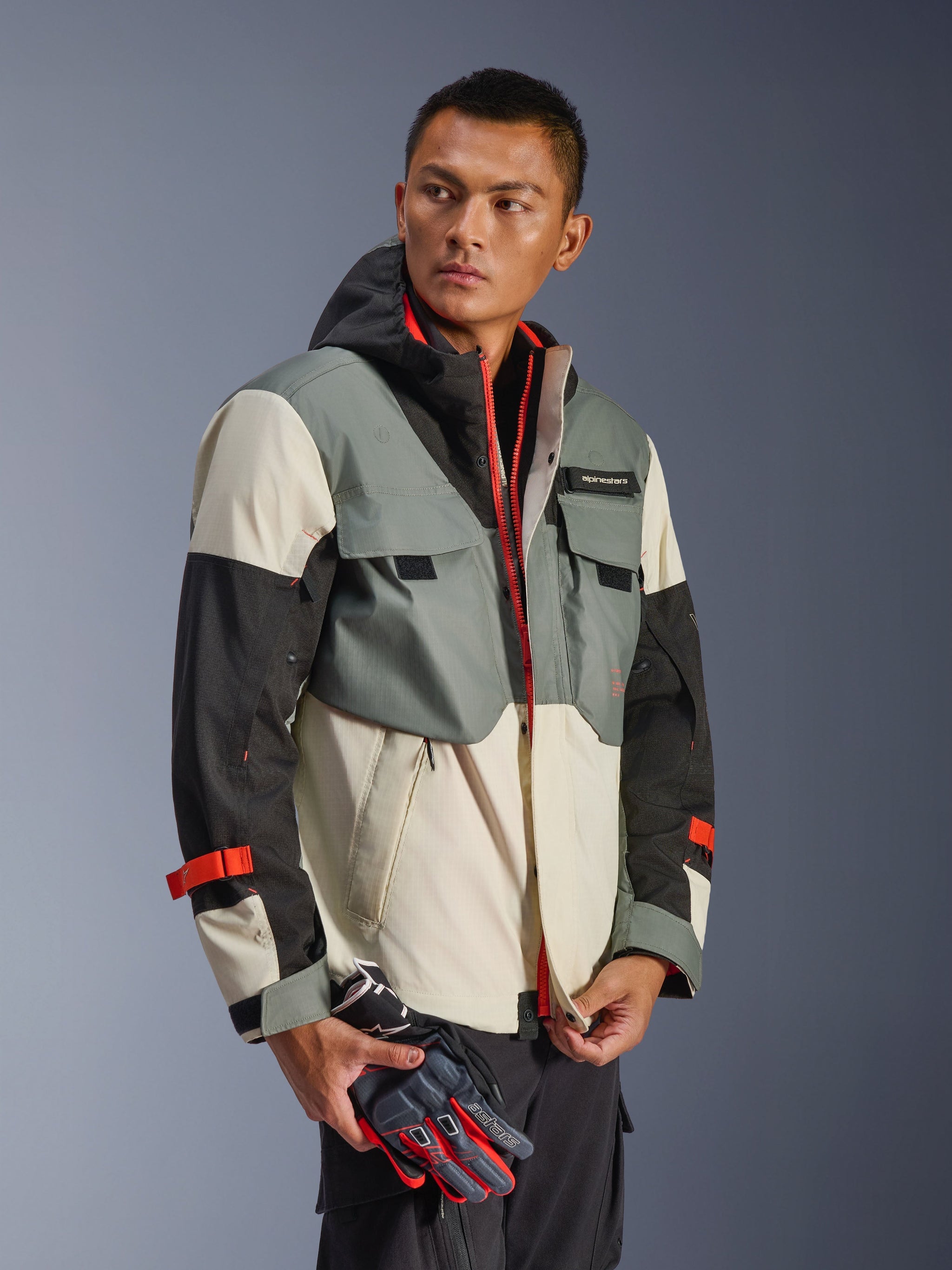 Alpinestars Mospher Waterproof Jacket in Fur Gray Black Ice, multi-panel textile parka with gray, black, and off-white sections, featuring bright red zipper and strap accents, tactical-inspired design with hood and utility pockets, worn by a model holding motorcycle gloves.