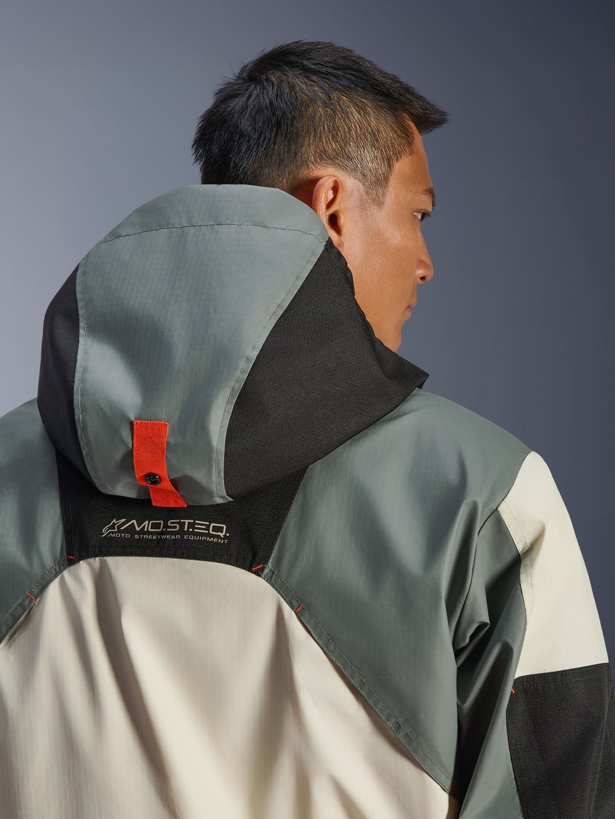 Alpinestars Mospher Waterproof Jacket, Fur Gray Black Ice, close-up back view showing the hood anchoring system with an orange snap button tab, MO.ST.EQ. Moto Streetwear Equipment branding, durable ripstop textile construction