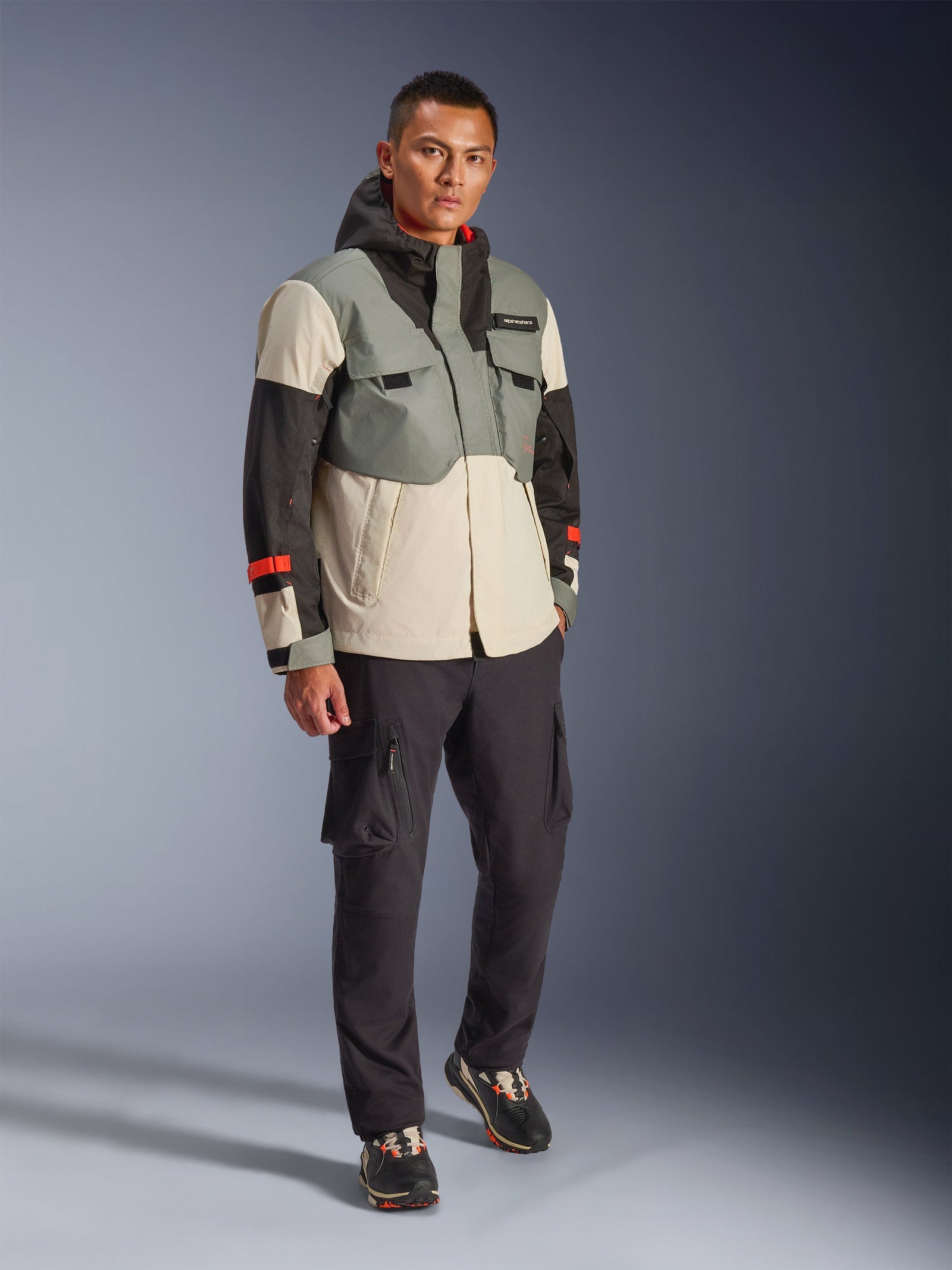 Alpinestars Mospher Waterproof Jacket in Fur Gray Black Ice, multi-colored technical motorcycle parka with gray, black, and off-white panels and red accents, utility-driven design with large pockets and hood, worn with dark gray cargo pants and black sneakers, urban contemporary motorcycle gear