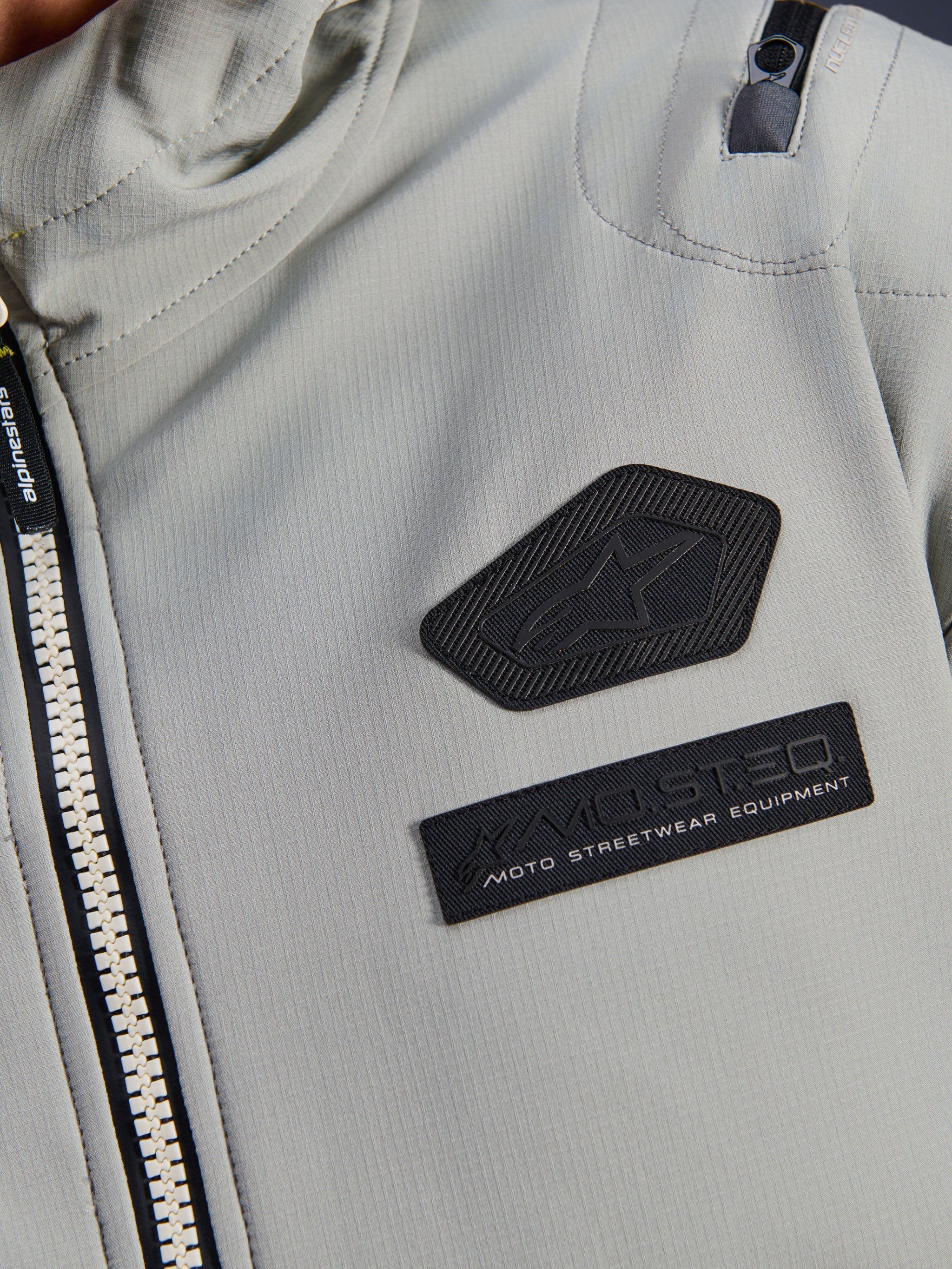 Alpinestars Moblast Waterproof Jacket, Iron Gray, close-up detail of chest patches featuring the Alpinestars logo and MO.ST.EQ. Moto Streetwear Equipment branding, technical ripstop fabric with heavy-duty front zipper and shoulder pocket detail.