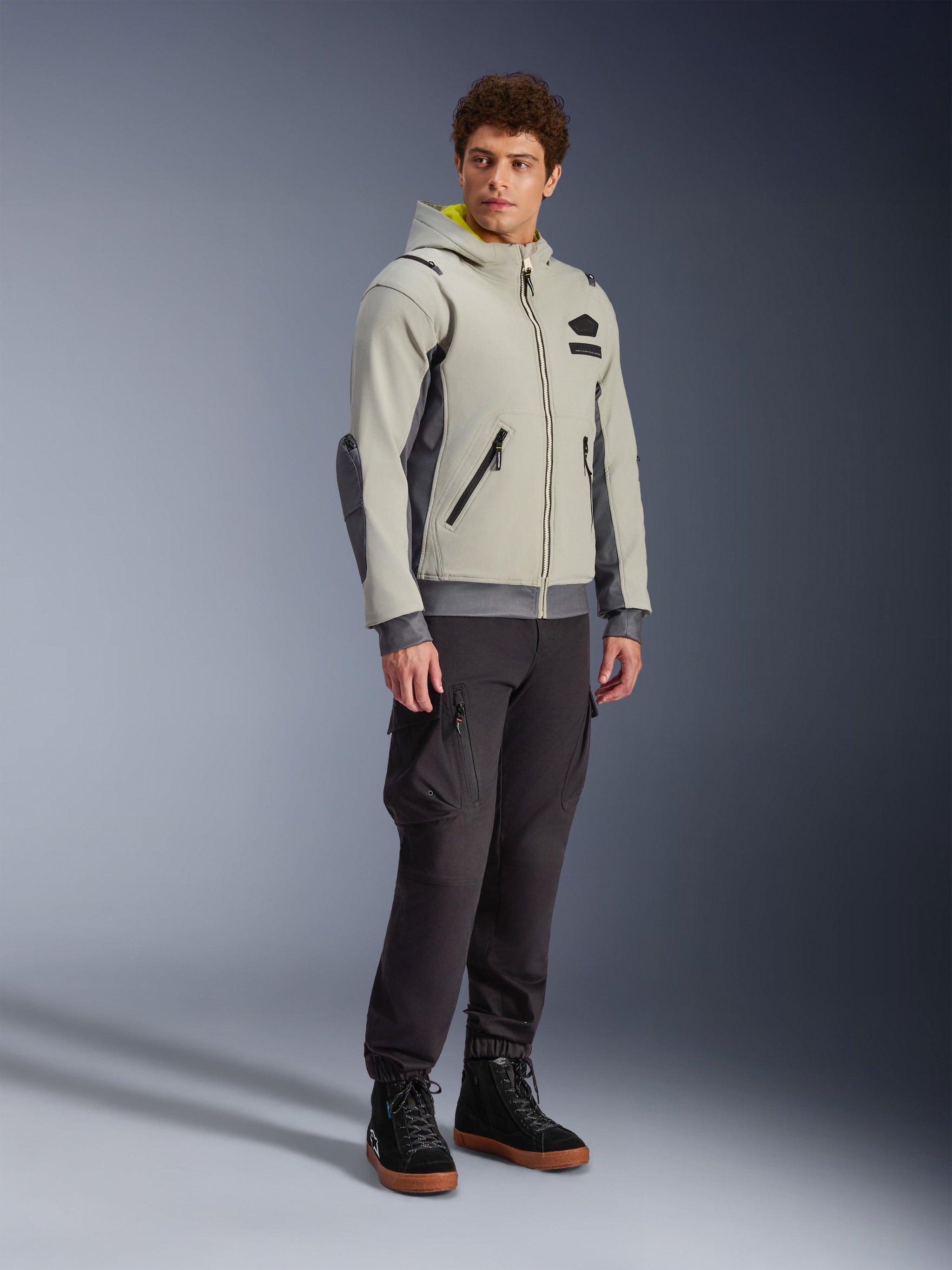 Alpinestars Moblast Waterproof Jacket, Iron Gray and Dark Sage, urban motorcycle riding jacket with a hoodie design, worn by a model with dark riding pants and black boots, minimalist cyber-tech aesthetic with a yellow interior hood and waterproof zippers