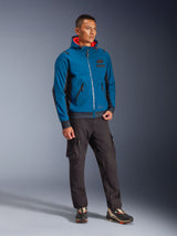 Male model wearing the Alpinestars Moblast Waterproof Jacket in Dark Petrol and Dark Navy, featuring a hooded design with a bright orange interior, paired with black cargo pants and sneakers, modern urban motorcycle riding gear.