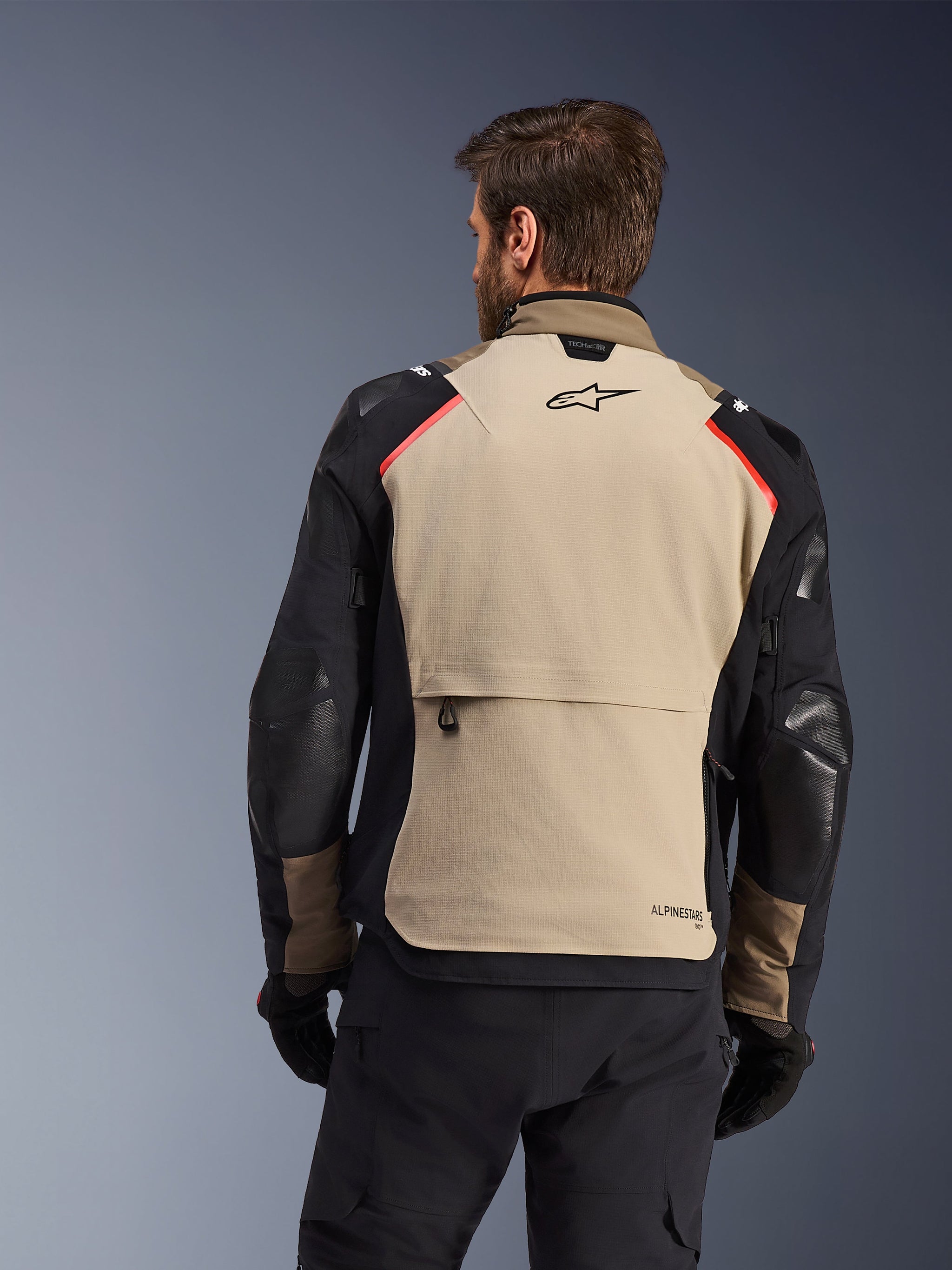 Alpinestars Andes Pro Drystar® XF Jacket, Rear View, Walnut and Aluminum, featuring a large tan back panel with black Alpinestars logo and red shoulder accents, large rear cargo pocket, worn with black pants and gloves, adventure touring motorcycle gear