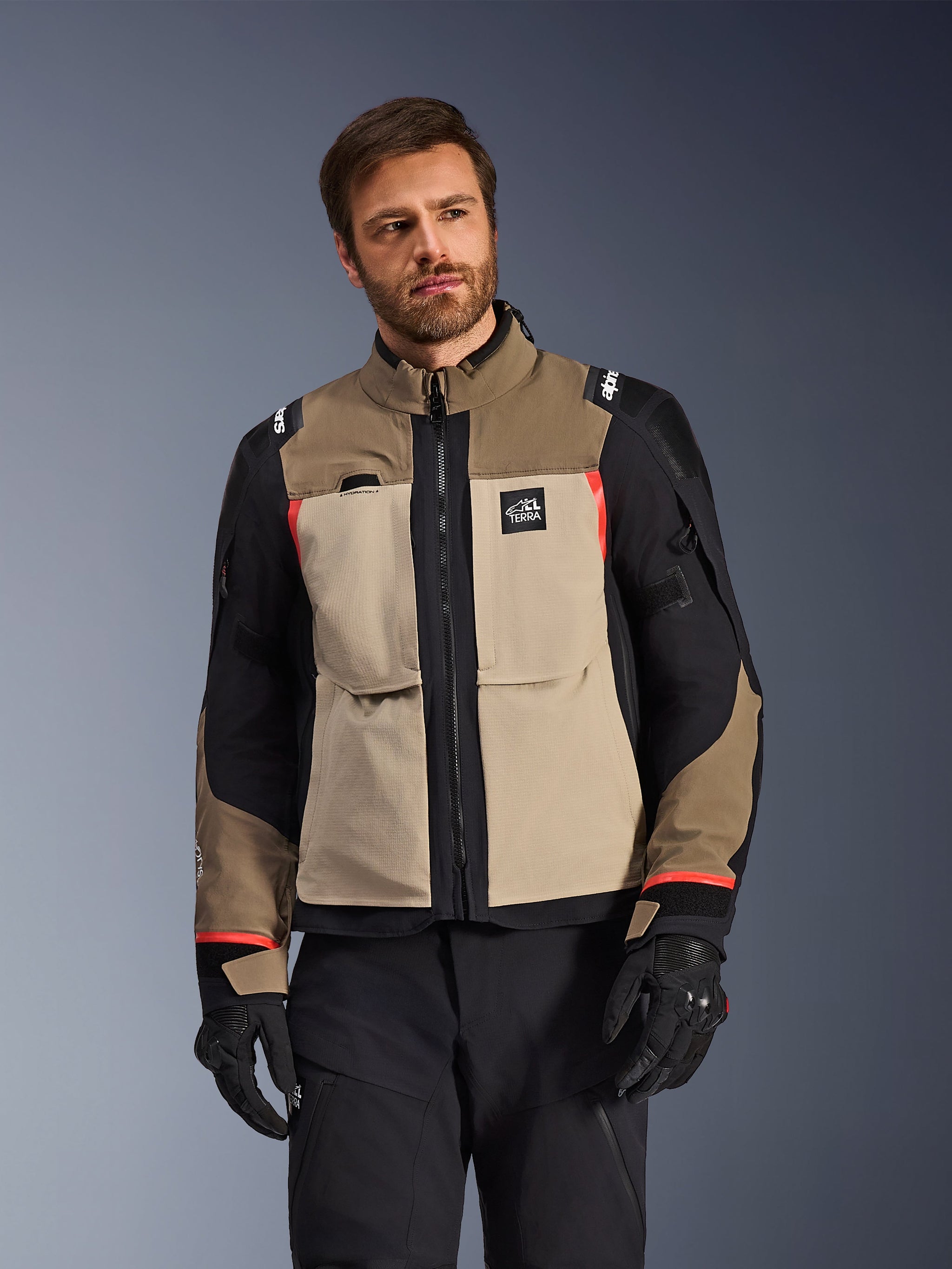 Alpinestars Andes Pro Drystar® XF Jacket, Walnut Aluminum, motorcycle adventure jacket featuring tan and black panels with red accents, worn with black riding pants and gloves, technical waterproof touring gear