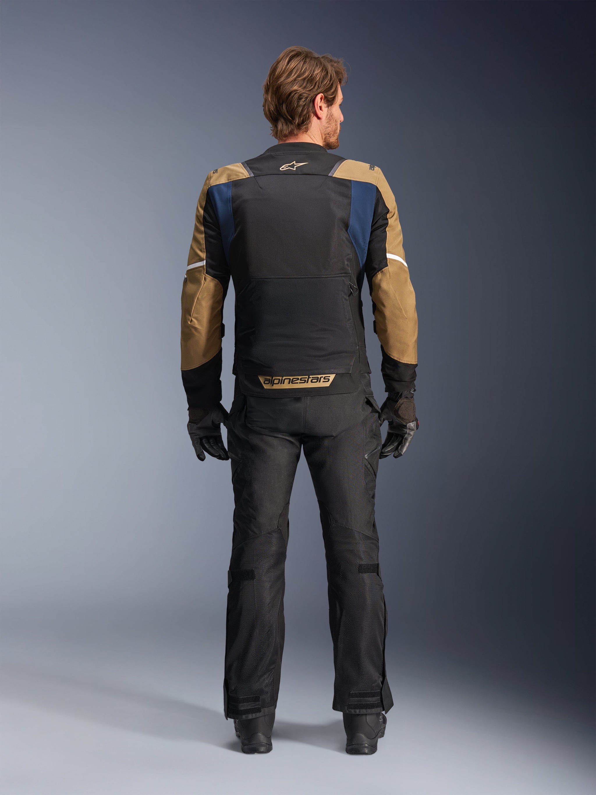 Alpinestars ST-2 Air Jacket in Deep Blue, Dark Khaki, and Black, rear view on model showing large mesh back panel and khaki sleeves with white stripes, paired with black technical motorcycle pants and gloves for adventure touring.