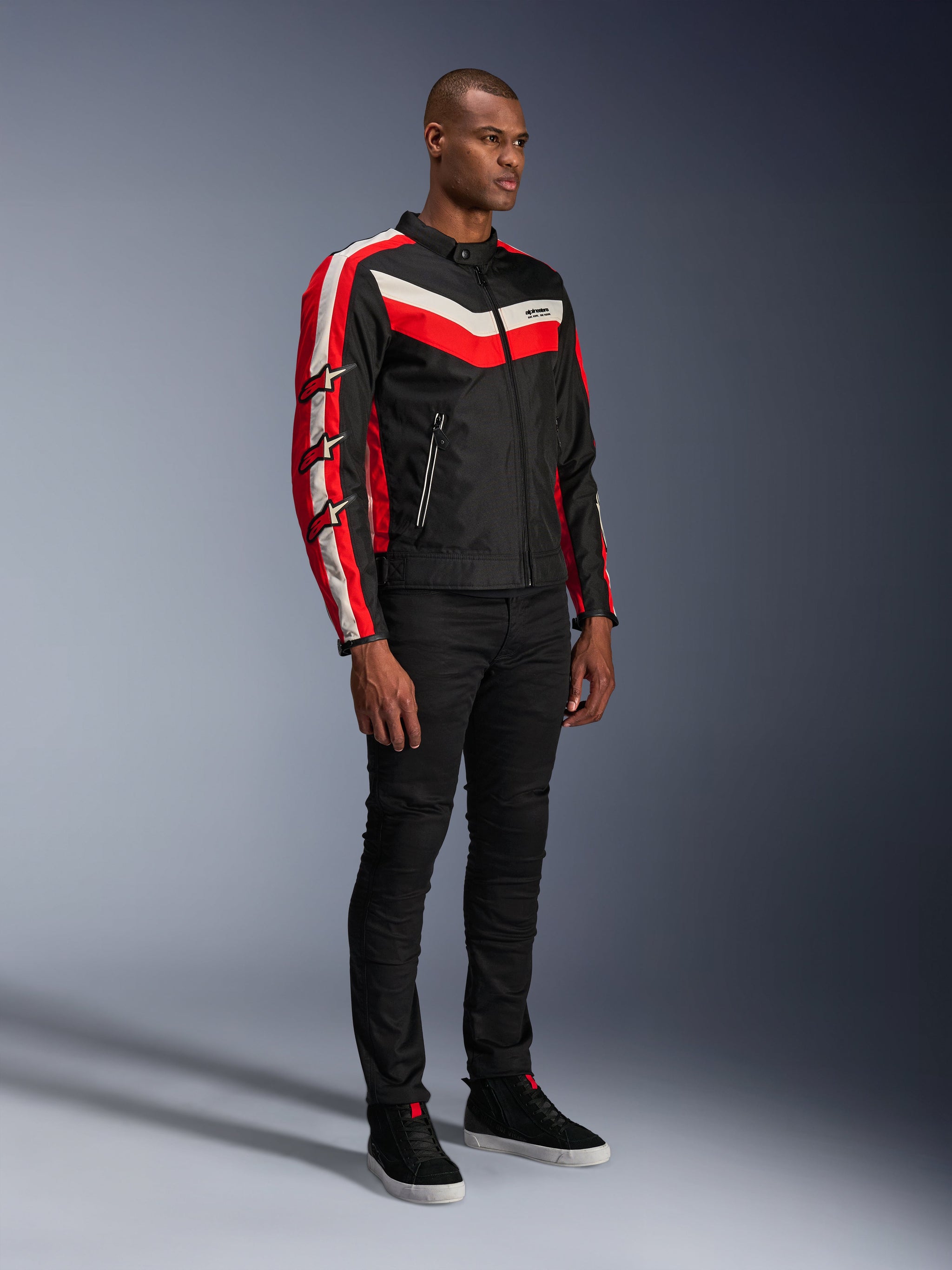 Male model wearing Alpinestars T-Dyno WR Jacket, textile motorcycle jacket in Black Bright Red Ecru, featuring red and off-white stripes and Alpinestars star logos on the sleeves, paired with black pants and black high-top sneakers, vintage-inspired urban riding gear.