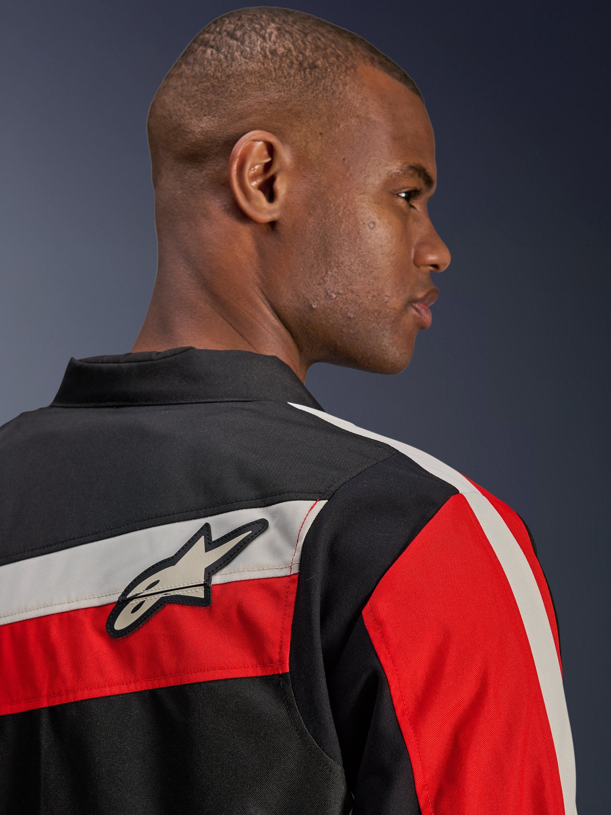 Alpinestars T-Dyno WR Jacket, close-up back view on a model, textile motorcycle jacket in black with horizontal ecru and bright red stripes, featuring the Alpinestars star logo patch on the shoulder, heritage-inspired biker style