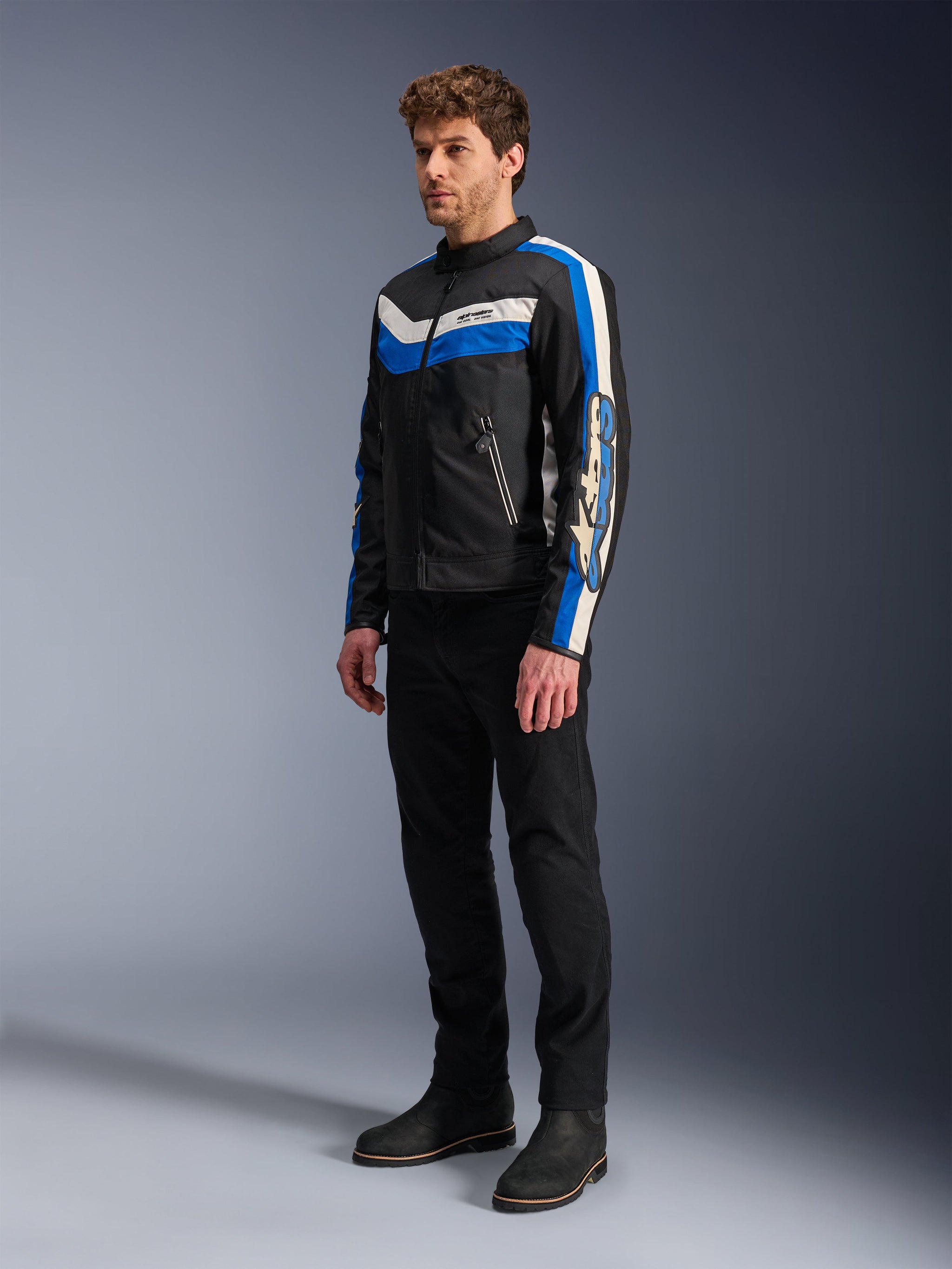 Alpinestars T-Dyno WR Jacket, Black Blue and Ecru, textile motorcycle jacket worn by model with black pants and boots, vintage-inspired design with bold blue and ecru stripes and branding on the sleeves, studio photography