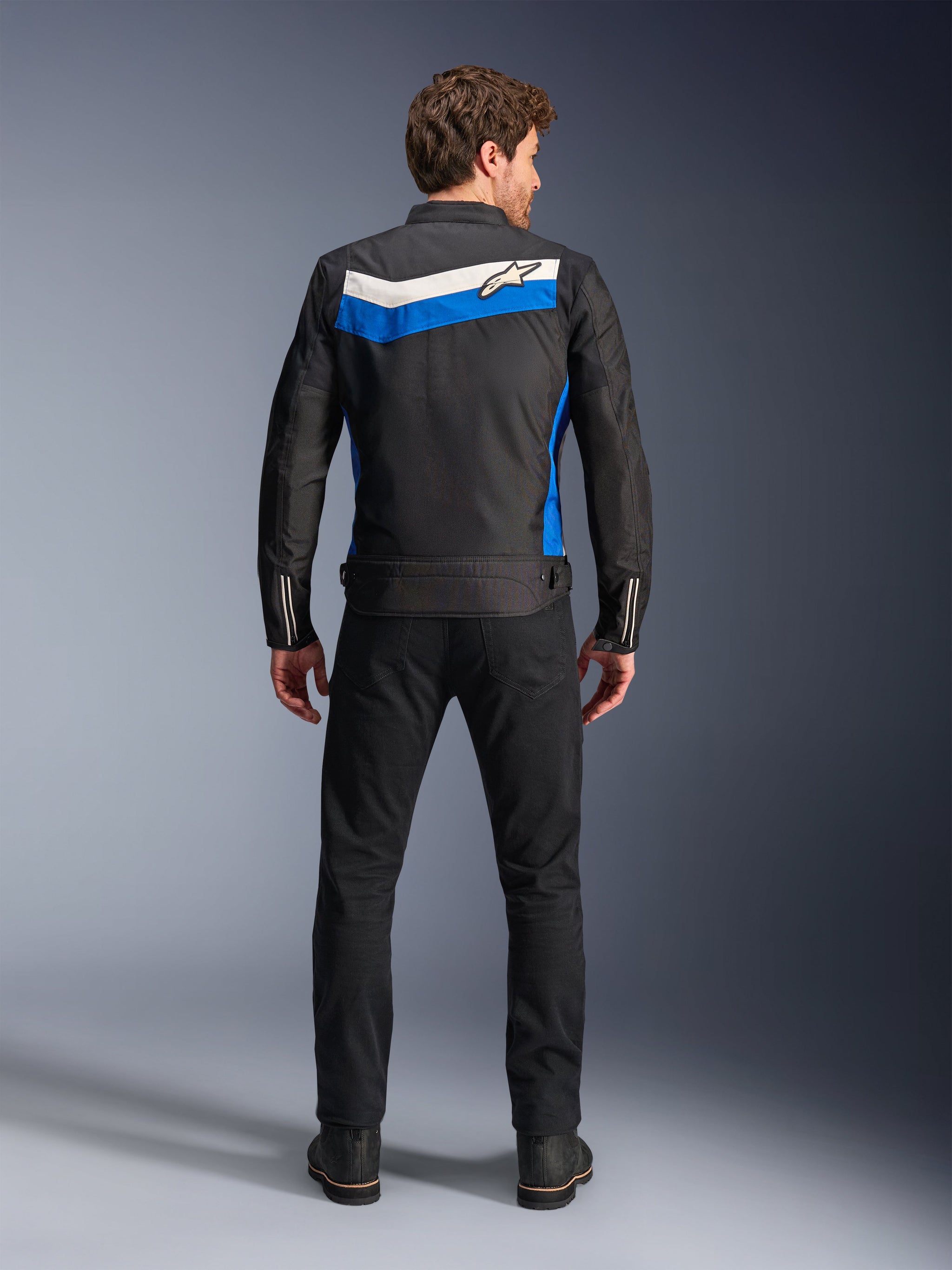 Alpinestars T-Dyno WR Jacket, textile motorcycle jacket, Black Blue Ecru colorway, full back view on model, featuring a blue and off-white chevron stripe with Alpinestars logo, paired with black pants and boots, vintage-inspired urban riding gear