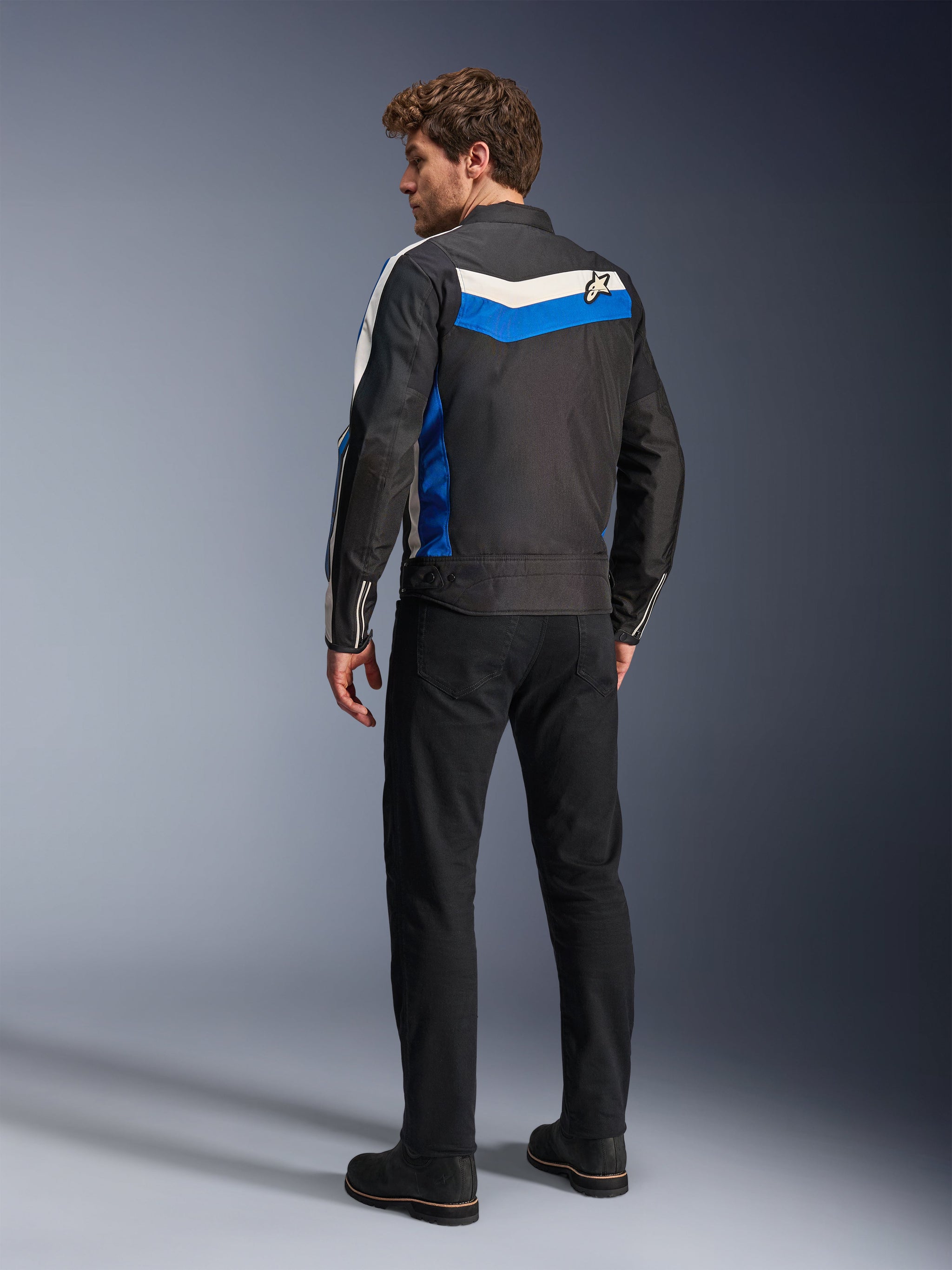 Alpinestars T-Dyno WR Jacket in Black Blue Ecru, back view on a male model, featuring a white and blue chevron stripe across the shoulders with a star logo, vintage-inspired textile motorcycle jacket, worn with black pants and boots