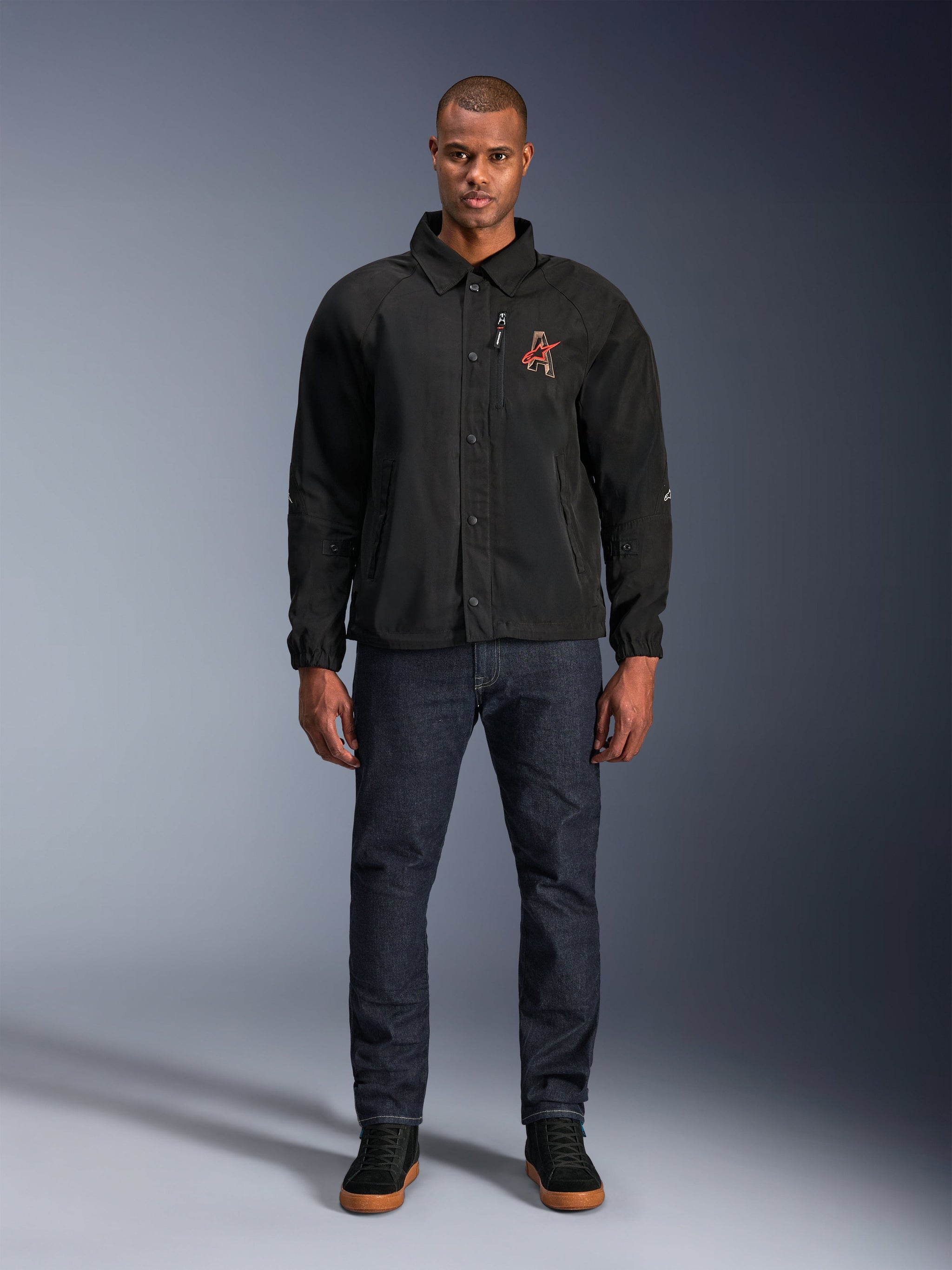 Alpinestars Revved WR Jacket in Black, worn by a model with dark blue jeans and black sneakers, front view, featuring snap button closure and a 3D embroidered logo on the chest, urban contemporary motorcycle gear.