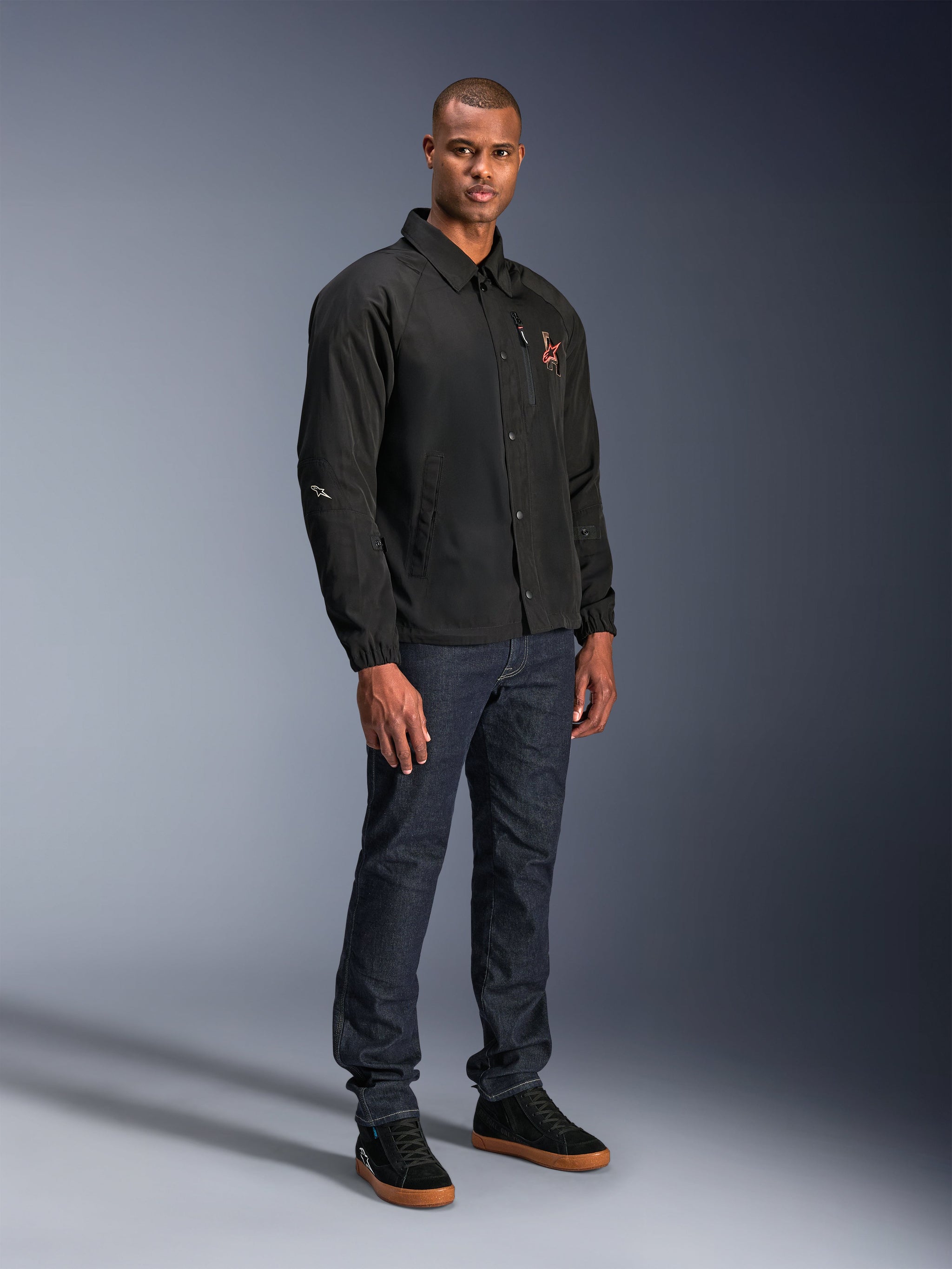 Alpinestars Revved WR Jacket in Black, urban coaches-style motorcycle jacket worn by a model with dark blue jeans and black sneakers, featuring a snap-button front and embroidered logo, minimalist and sporty design for road riding.