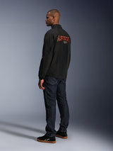 Alpinestars Revved WR Jacket, Black, featuring large orange 3D embroidered Alpinestars branding on the back, rear three-quarter view of a model wearing dark blue jeans and black sneakers, urban contemporary motorcycle jacket with a coaches-jacket silhouette