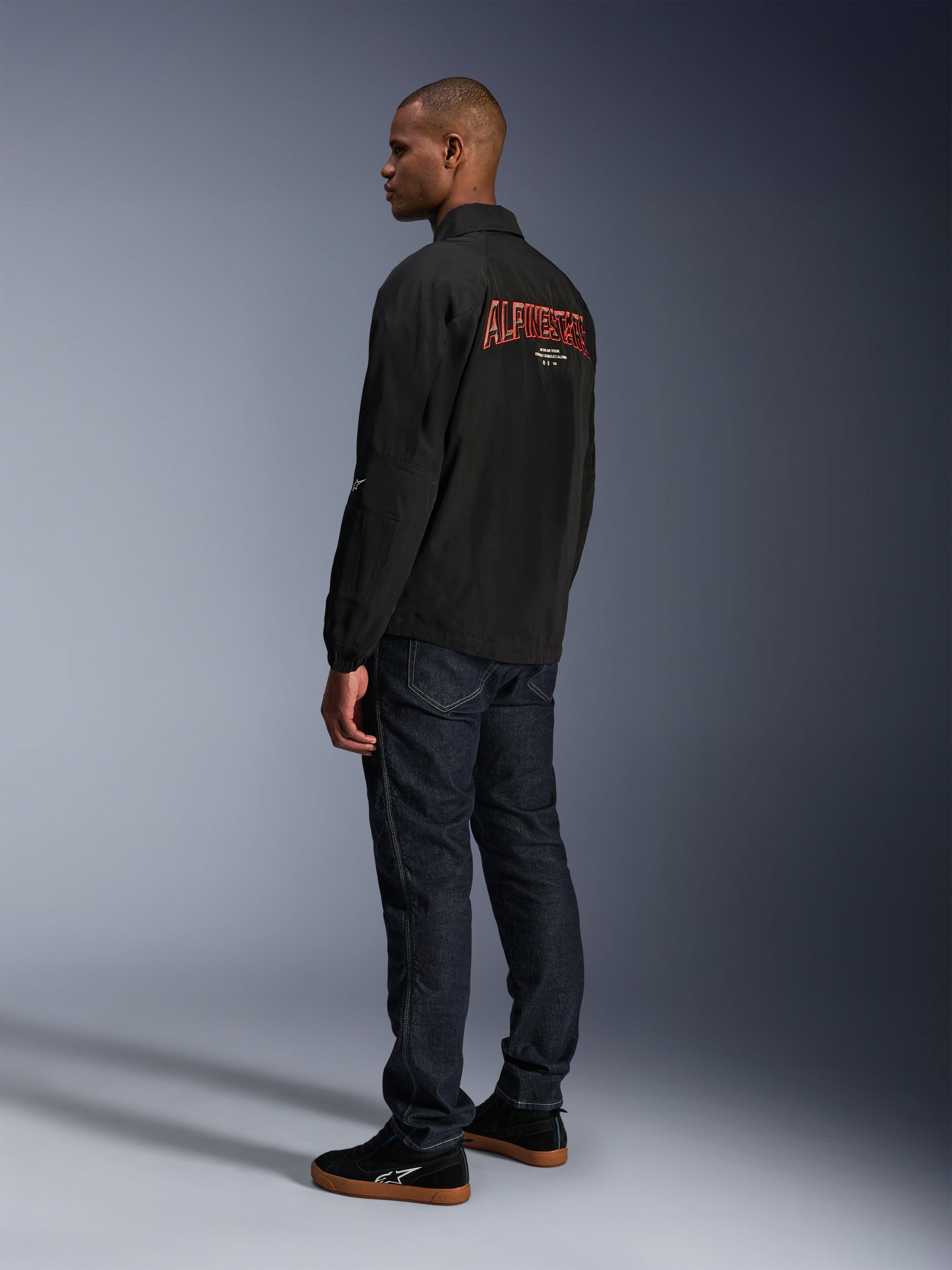 Alpinestars Revved WR Jacket, Black, featuring large orange 3D embroidered Alpinestars branding on the back, rear three-quarter view of a model wearing dark blue jeans and black sneakers, urban contemporary motorcycle jacket with a coaches-jacket silhouette