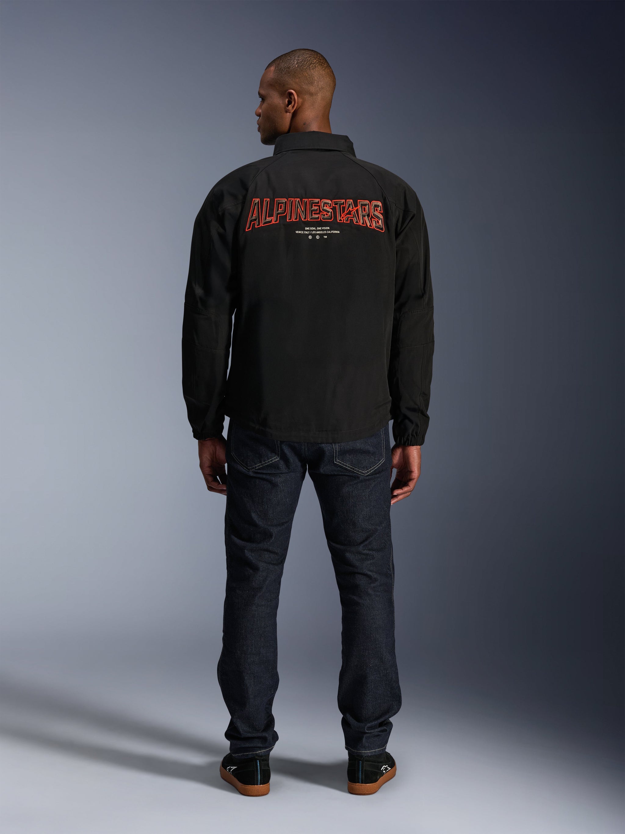 Alpinestars Revved WR Jacket, Black, back view featuring large red 3D-effect 'ALPINESTARS' embroidery, urban coaches-style jacket, worn with dark denim jeans and black sneakers, minimalist motorcycle apparel