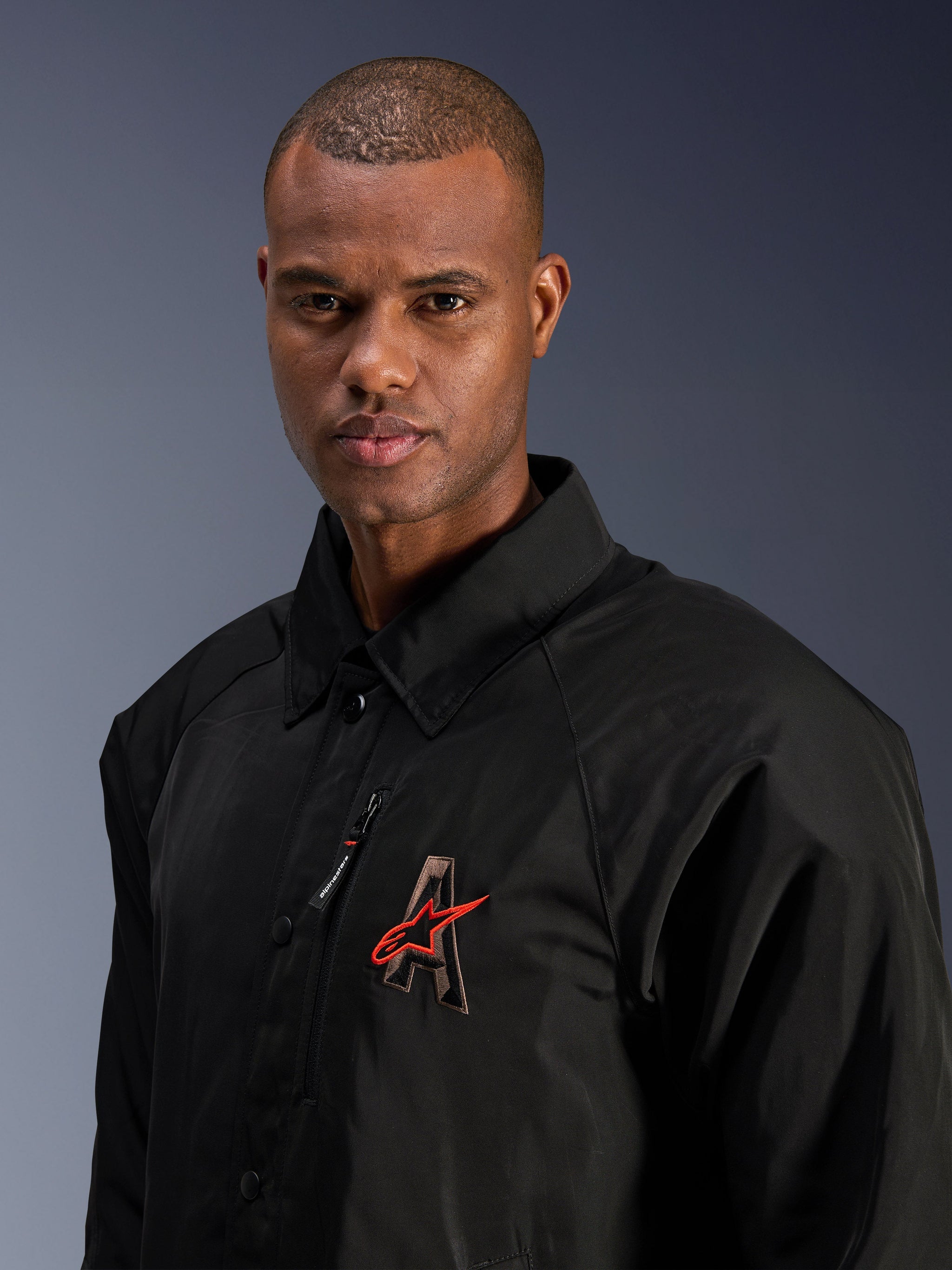 Alpinestars Revved WR Jacket, Black, close-up shot of a man wearing the water-resistant satin motorcycle jacket, featuring 3D embroidered 'A' logo with red Alpinestars star, zippered chest pocket, and collared design for urban contemporary riding.