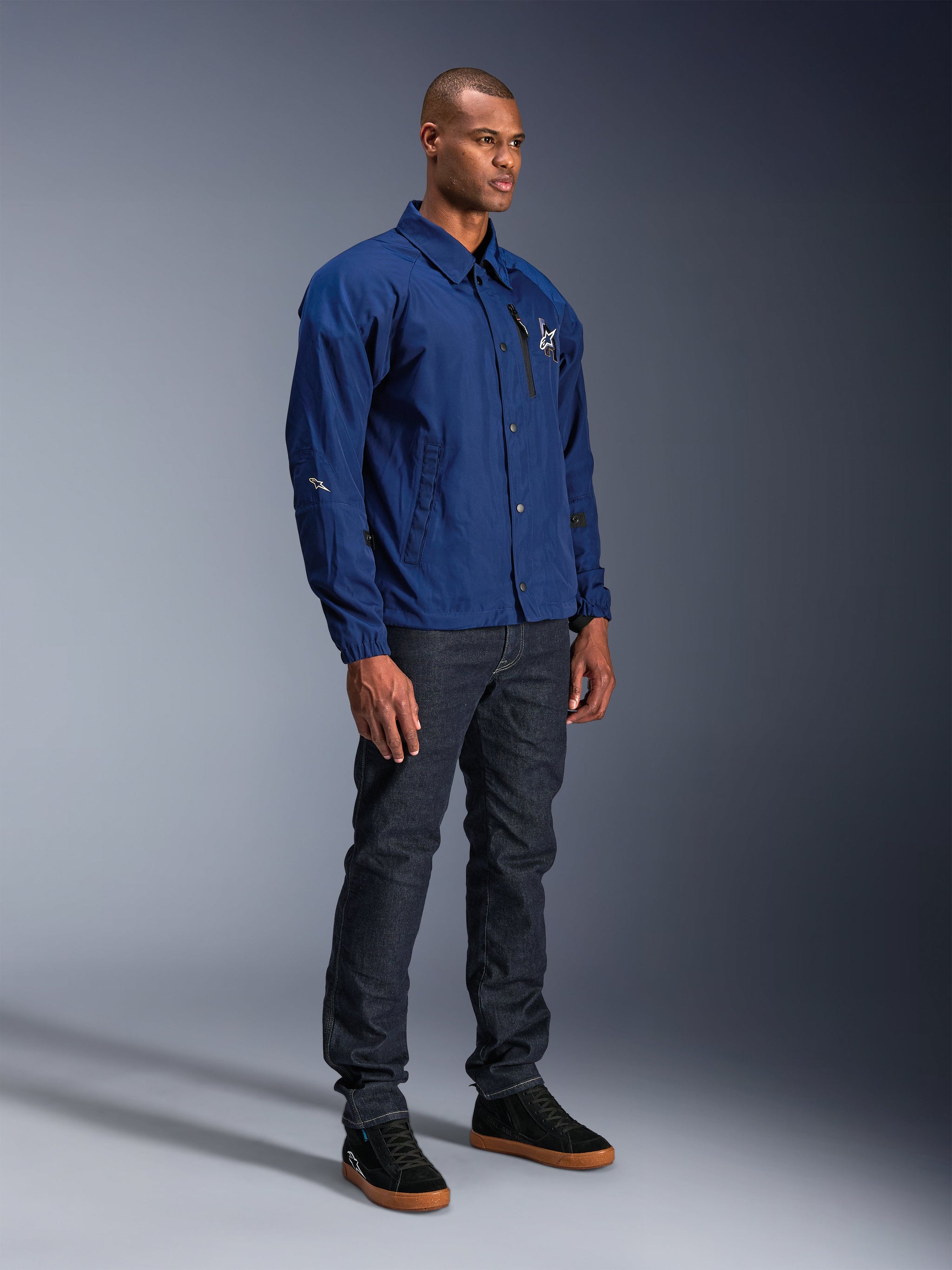 Alpinestars Revved WR Jacket, Blue motorcycle jacket, worn by male model with dark denim jeans and black sneakers, featuring a water-resistant coaches jacket silhouette with snap button front and minimalist chest logo.