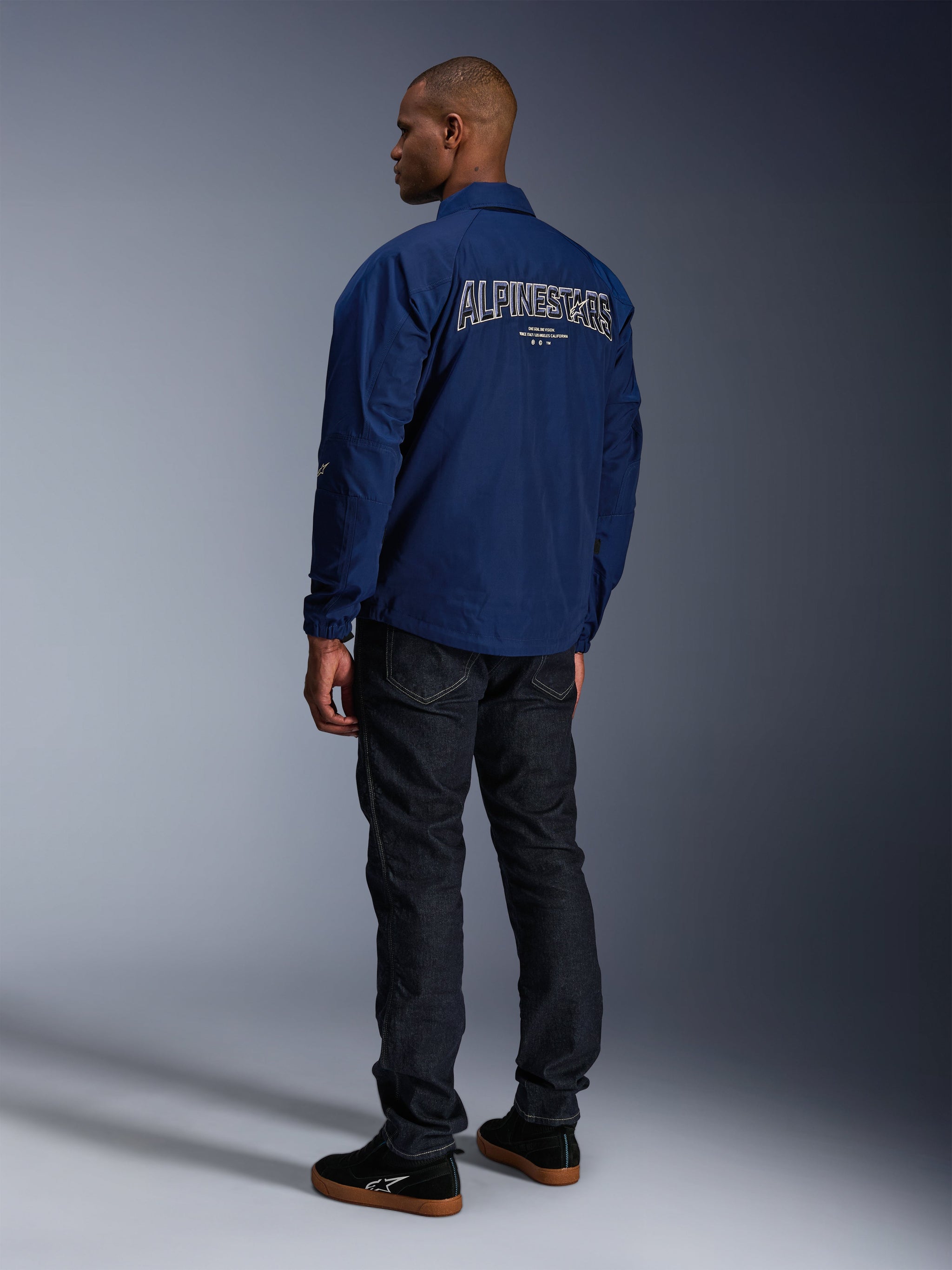Model wearing the Alpinestars Revved WR Jacket in Navy Blue from behind, featuring large white outlined 'ALPINESTARS' embroidery across the back, paired with dark denim pants and black sneakers, urban motorcycle aesthetic.