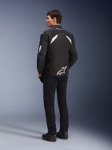 Male model from the back wearing the Alpinestars T-GP R V3 Drystar® Jacket in Black and White, featuring white shoulder accents and a large white logo on the lower back, paired with black riding pants and boots, sport textile motorcycle jacket.