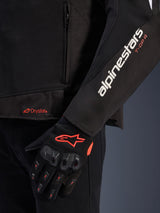 Alpinestars T-GP R V3 Drystar® Jacket, black textile motorcycle jacket, close-up view showing white alpinestars and orange T-GP R sleeve branding, orange Drystar logo on the hem, paired with black protective mesh gloves with red accents.