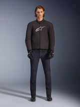 Model wearing Alpinestars SMX Waterproof Jacket in Black with white and red logo, paired with dark denim riding pants, black gloves, and riding boots, sporty all-weather motorcycle gear outfit.