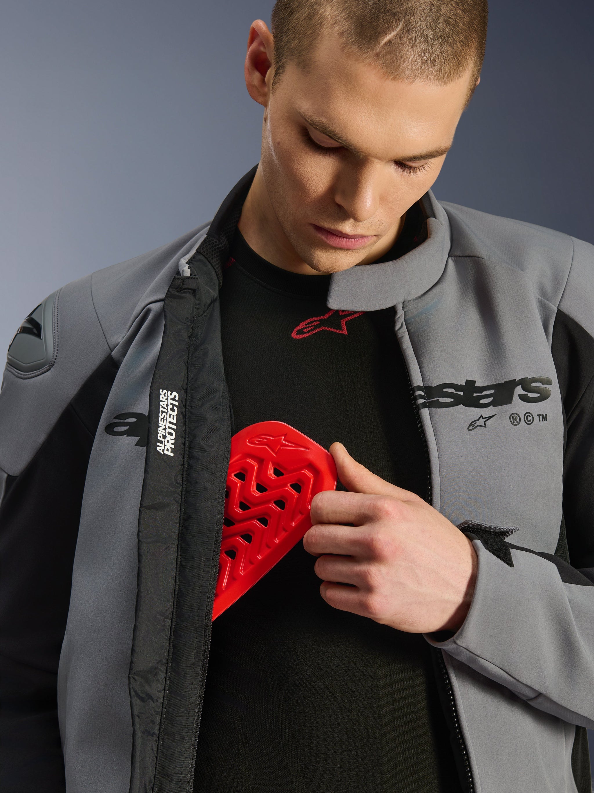 Male model wearing the Alpinestars SMX Waterproof Jacket in Dark Gray Black, inserting a red Nucleon chest protector into an internal pocket, showcasing the jacket's armor compatibility and protective features.