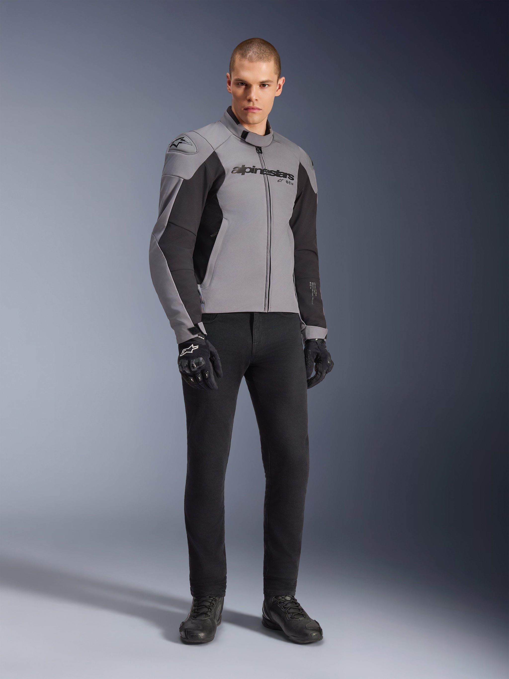 Alpinestars SMX Waterproof Jacket, Motorcycle Jacket, Dark Gray and Black, worn by model with black riding gloves and black pants, featuring bold chest branding and a technical sport-mapped design for all-weather road riding.