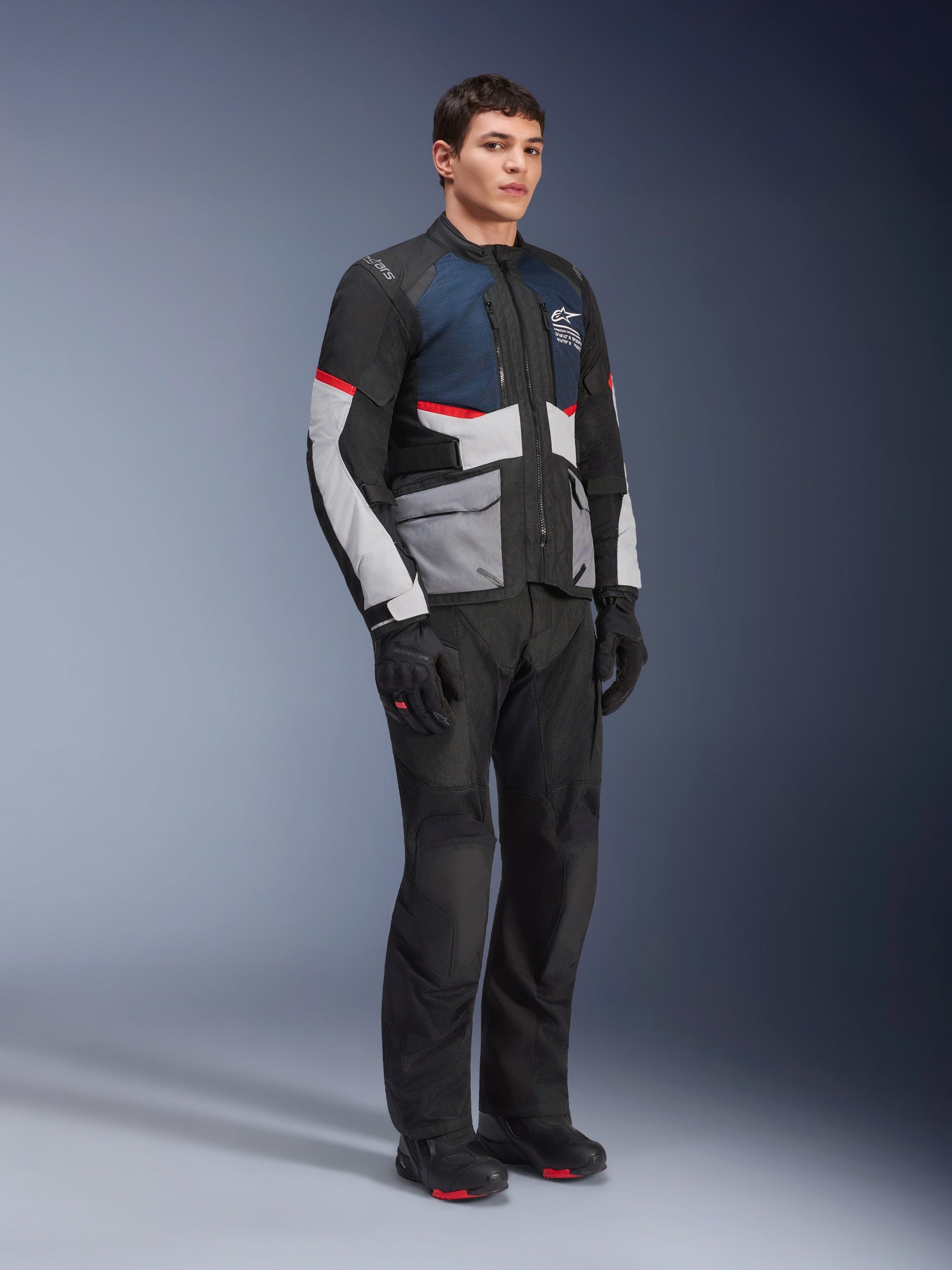Male model wearing the Alpinestars Andes Air Drystar® Jacket in Deep Blue Black Ice Gray, paired with black motorcycle pants, gloves, and boots, featuring large mesh panels for ventilation, light gray pockets, and red accents, designed for adventure touring.