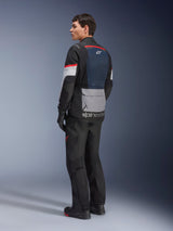 Male model from a rear-side view wearing the Alpinestars Andes Air Drystar® Jacket in Deep Blue Black Ice Gray, paired with black motorcycle pants and gloves, featuring a blue mesh back panel, gray utility pocket on the lower back, and red accents on the sleeves, professional adventure touring gear.