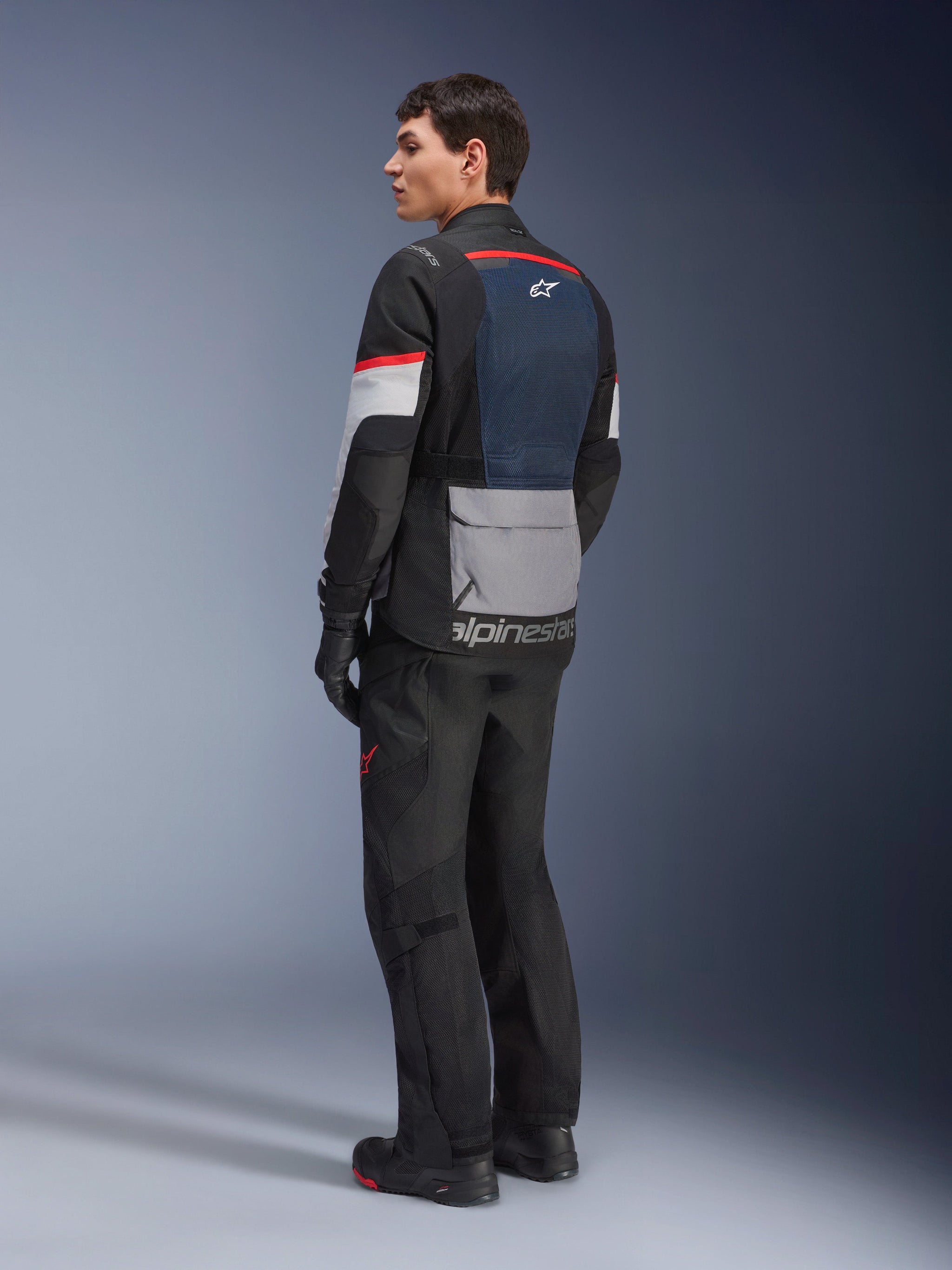 Male model from a rear-side view wearing the Alpinestars Andes Air Drystar® Jacket in Deep Blue Black Ice Gray, paired with black motorcycle pants and gloves, featuring a blue mesh back panel, gray utility pocket on the lower back, and red accents on the sleeves, professional adventure touring gear.
