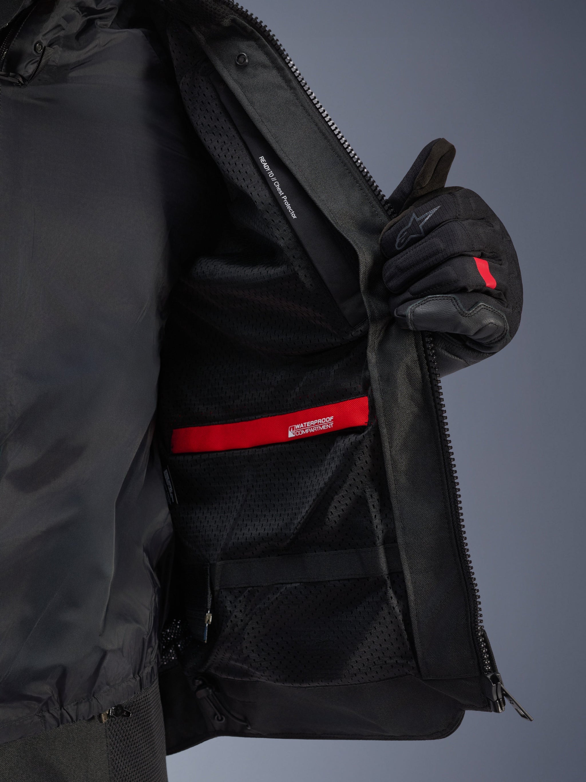 Interior detail view of the Alpinestars Andes Air Drystar® Jacket, highlighting the breathable black mesh lining, red waterproof compartment, and chest protector pocket, shown being held open by a black motorcycle glove.