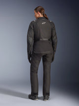 Female rider from behind wearing the Alpinestars Stella Andes V4 Drystar® Jacket, Black, paired with matching motorcycle pants, white Astar logo on the back, elongated rear hem, complete adventure-touring riding outfit including black gloves and boots