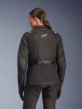 Woman wearing Alpinestars Stella Andes V4 Drystar Jacket in Black, rear view featuring white logo on the back and reinforced elbow panels, paired with matching black pants and gloves, adventure touring motorcycle outfit