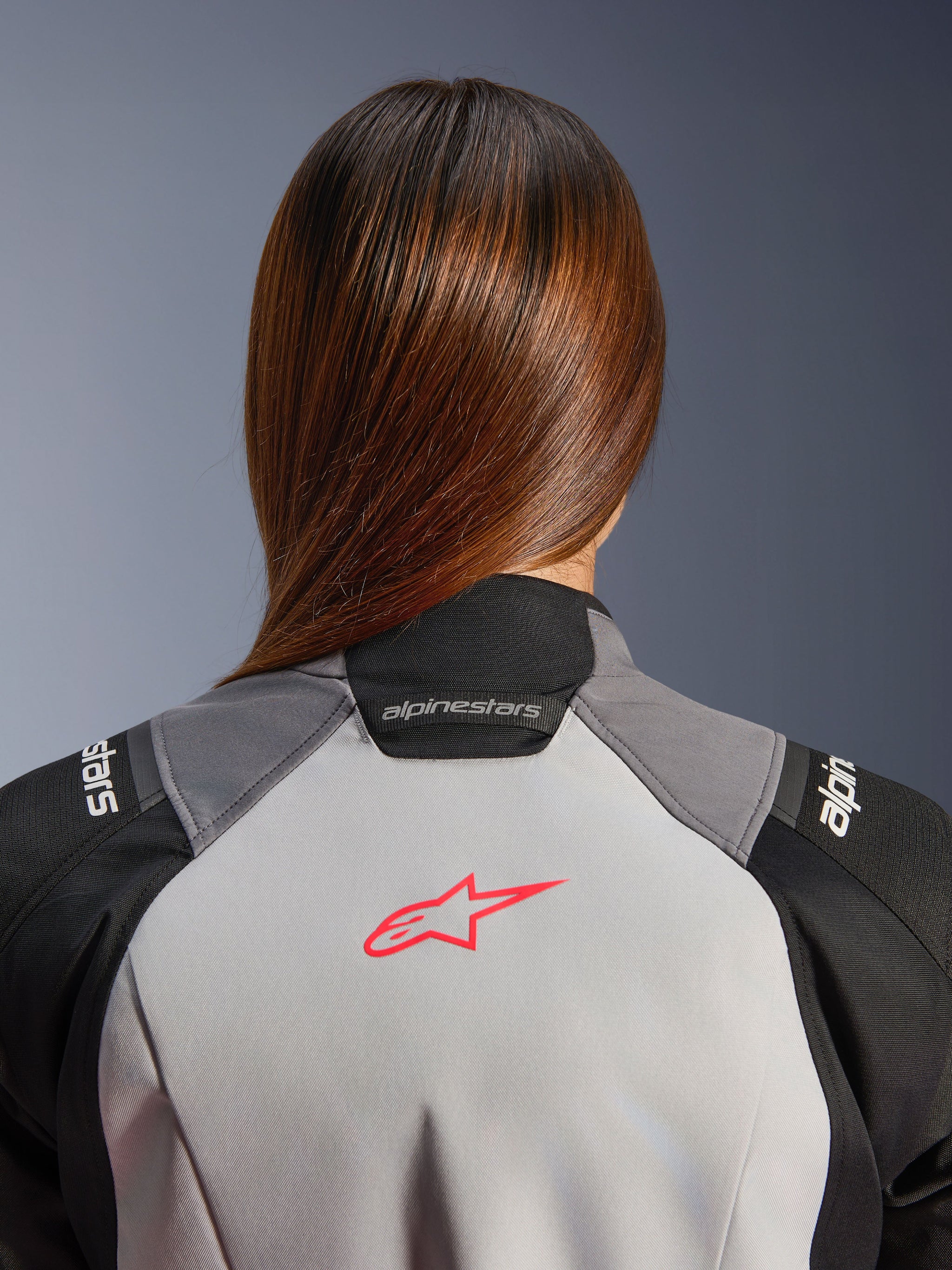 Alpinestars Woman Stella Andes V4 Drystar® Jacket, close-up of the upper back and collar, ice gray textile with a coral fluo star logo, black and dark gray panels, worn by a rider with long brown hair.